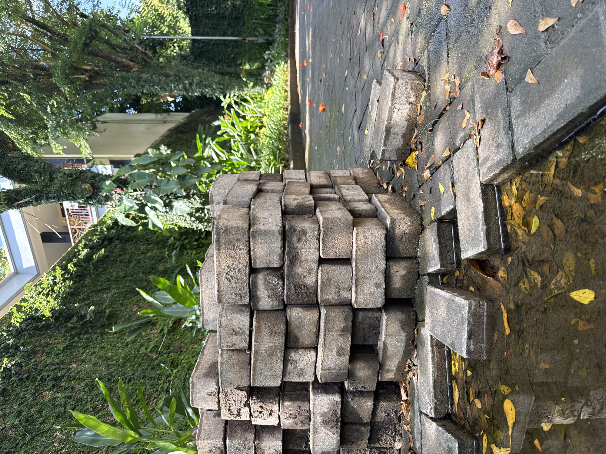 A vertical-oriented photograph shows a stack of gray bricks in an outdoor setting. The bricks are arranged in an organized manner, forming a rectangular block. The bricks appear to be made of stone and are weathered.
To the left of the bricks, there is a green, dense vegetation, possibly a wall covered in climbing plants. A building is barely visible behind the vegetation. To the right of the bricks, the ground is paved with more gray bricks. Some leaves litter the pavement. A small puddle of water is on the right side of the pavement.
Based on the information provided, this photo was taken in Jimbaran, Indonesia. However, without additional context or information, the exact location within Jimbaran cannot be determined. The absence of people, actions, and interactions makes it difficult to establish context beyond the physical elements in the frame.