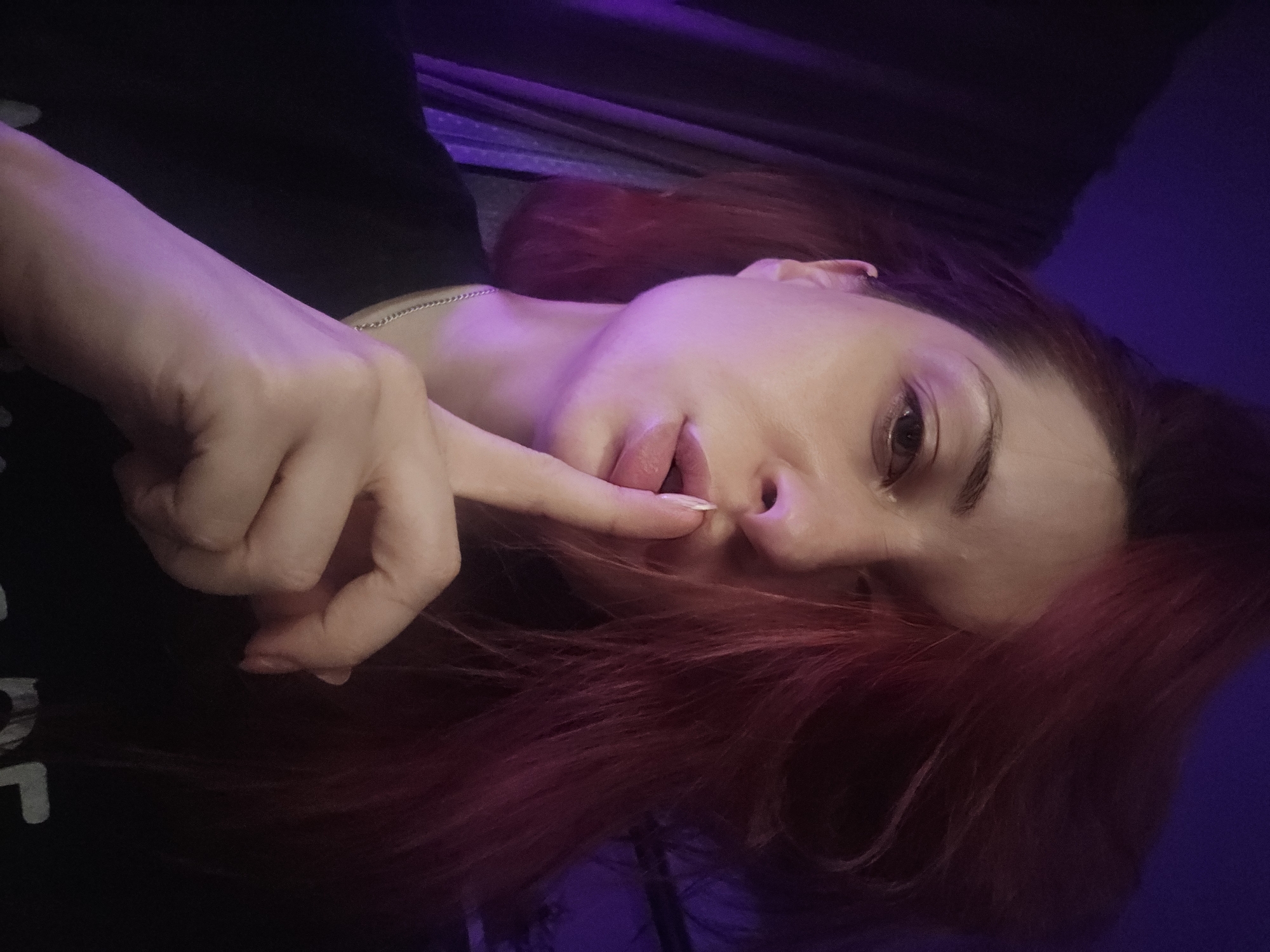 The image is a close-up, first-person perspective selfie of a woman with reddish-pink hair. She is wearing a black t-shirt and a silver chain necklace. Her right hand is raised to her lips, with her index finger pressed against them in a "shushing" gesture. Her eyes are looking towards the camera. The background is dimly lit with a purple hue, with what appears to be a dark curtain on the left side. The image does not contain any discernible information about the location in Burgas, Bulgaria.
