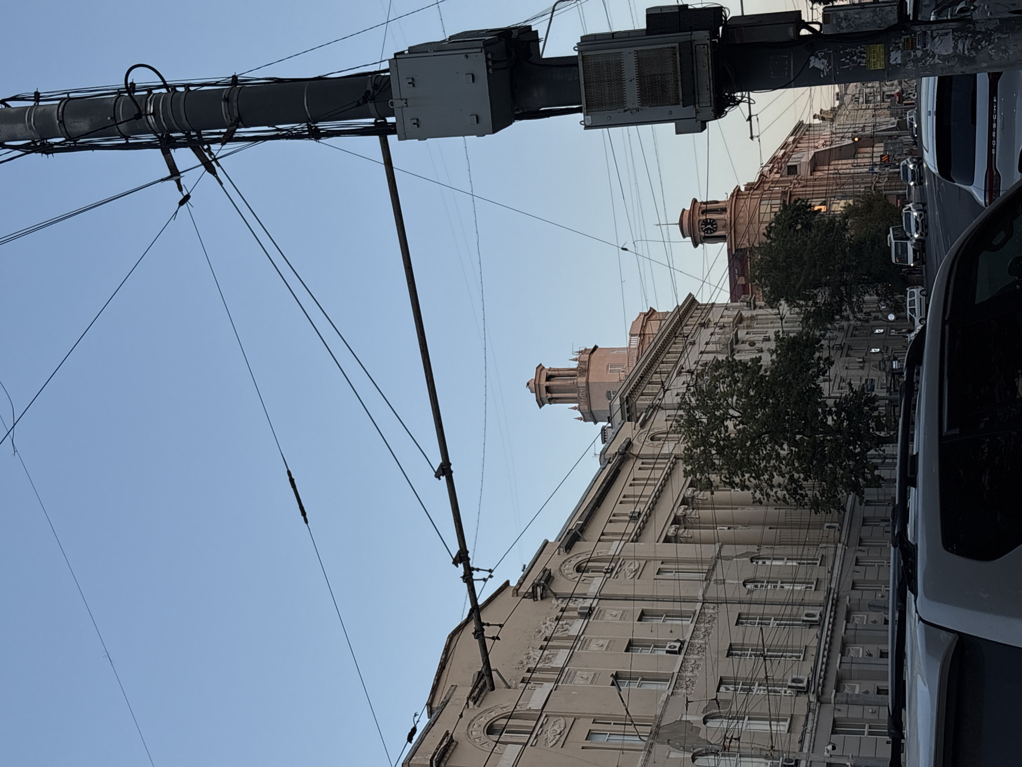 The image shows a street scene in Rostov-on-Don, Russia, likely during the daytime. In the foreground, the roof of a white SUV is visible. The street is lined with multi-story buildings, appearing to be of historical architecture, with detailed facades and large windows. Trees are interspersed along the buildings. A prominent utility pole with numerous wires and electrical boxes stands on the right side of the image. Several cars are driving on the street, including a white van and other sedans. In the background, two towers are visible atop the buildings; one has a clock face. The sky is clear and light blue. The overall impression is that of a busy urban street.