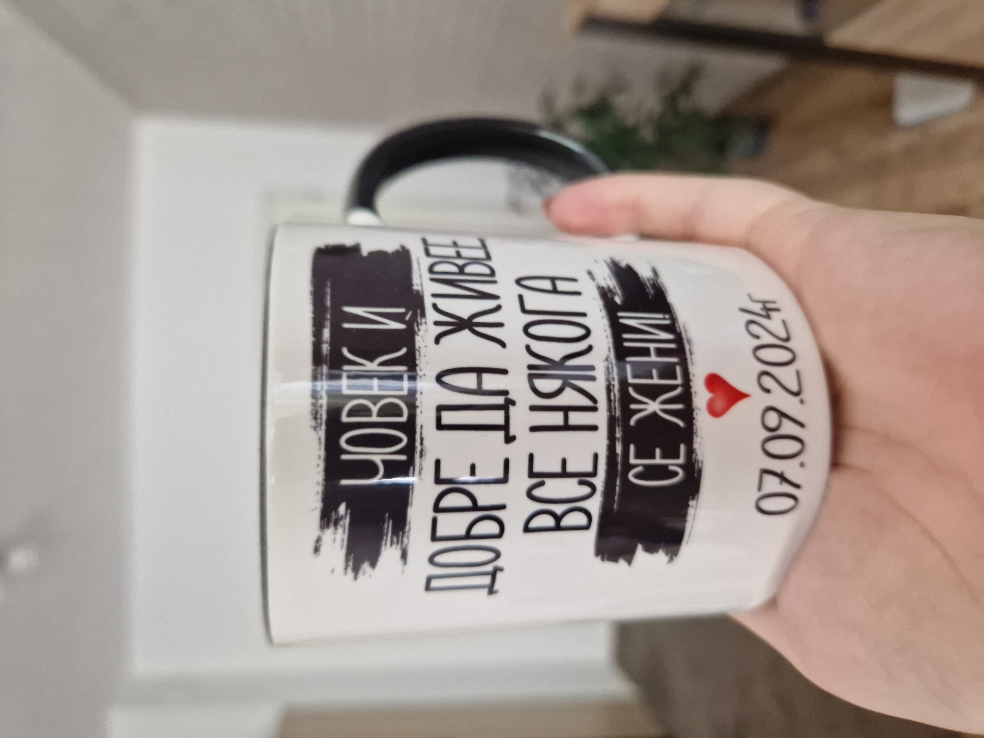A white ceramic mug with a black handle is centered, held by a light-skinned hand. The mug displays black Cyrillic text in a casual font. The text reads: "ЧОВЕК И ДОБРЕ ДА ЖИВЕЕ ВСЕ НЯКОГА СЕ ЖЕНИ!" which translates from Bulgarian as "A man, even if he lives well, will always somehow get married!". Some lines of text are superimposed on black, distressed horizontal bar graphics. Below the text, a small red heart symbol is positioned above the date "07.09.2024г". The hand supports the mug from its base. The background is an out-of-focus indoor environment, with light walls or structures visible to the left and a darker, blurred object (possibly a plant) against a lighter, textured wall to the right.