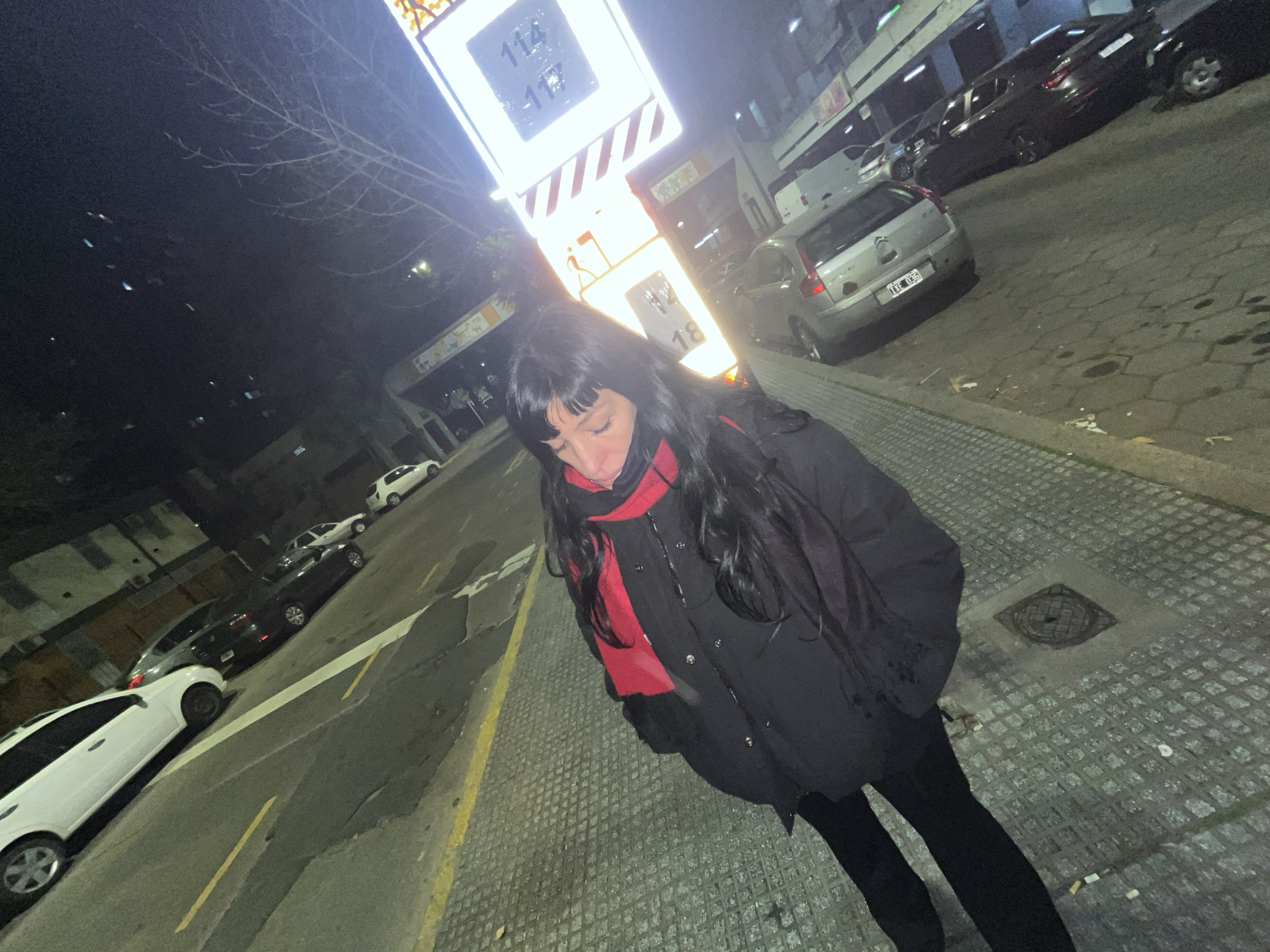 The image depicts a woman standing on a sidewalk at night. She is wearing a black coat and a red scarf. Behind her is a gas station sign showing gas prices. Cars are parked along the street. The location is most likely in Buenos Aires, Argentina, given the street style and car models.