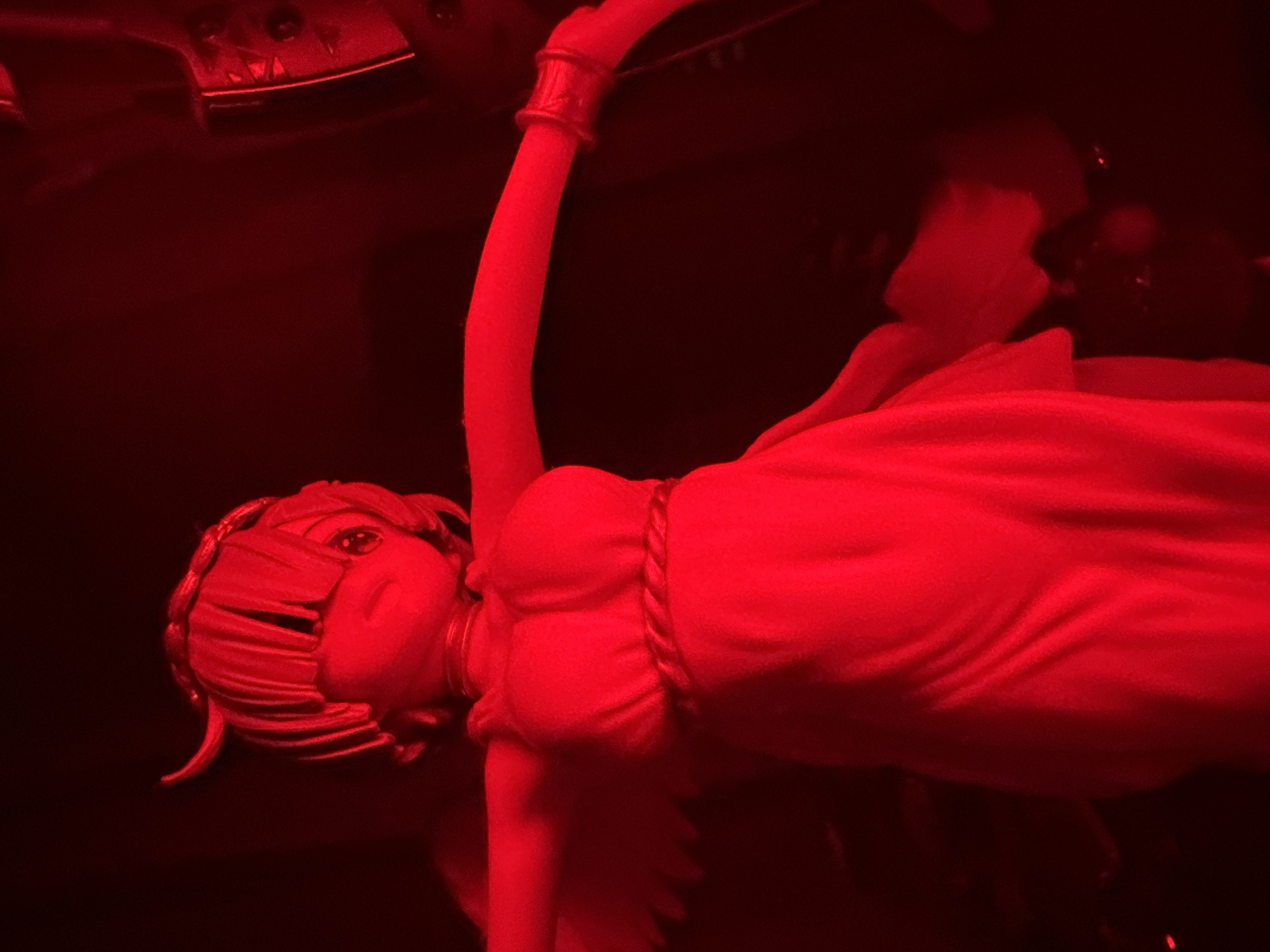 A figurine of a stylized female character, illuminated by a vibrant red light, stands with arms outstretched. The figurine has large, cartoonish eyes, short, dark hair that appears to be styled in bangs, and small, curved horns protruding from its head. It wears a flowing, draped dress with a braided belt around the waist and a circular adornment on its head. A winged silhouette is visible on its back. The figurine is positioned against a dark, indistinct background, with some blurred objects and reflections suggesting an indoor environment.