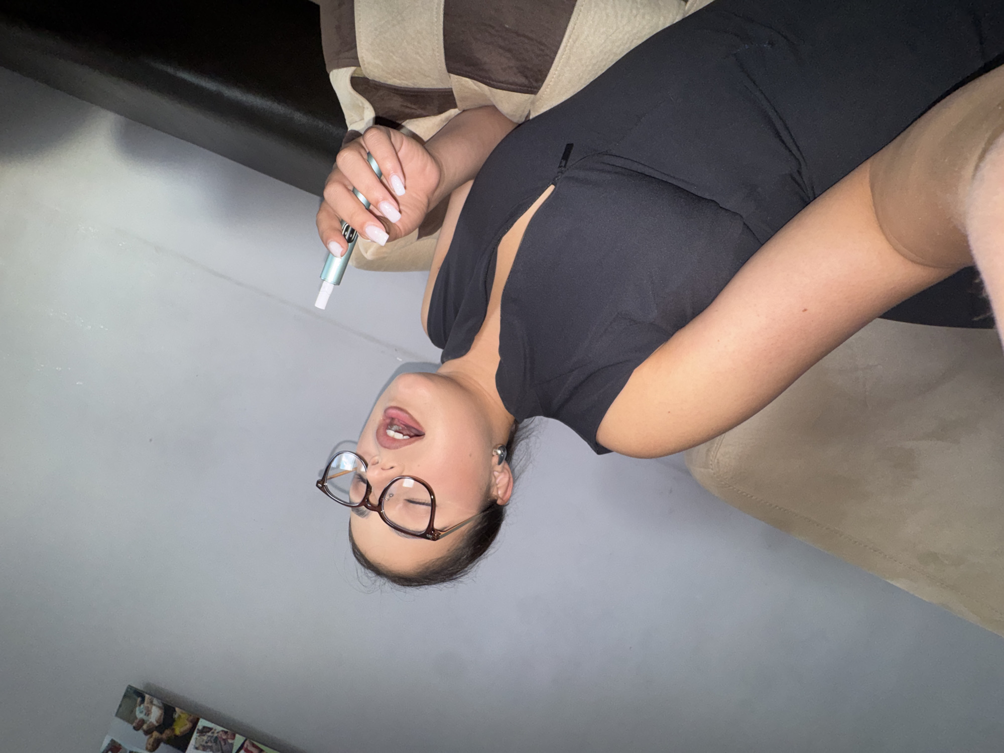 A woman with glasses is seen lying on a couch. She is wearing a black top and has an electronic cigarette in her hand. The setting appears to be indoors, possibly a residential space.