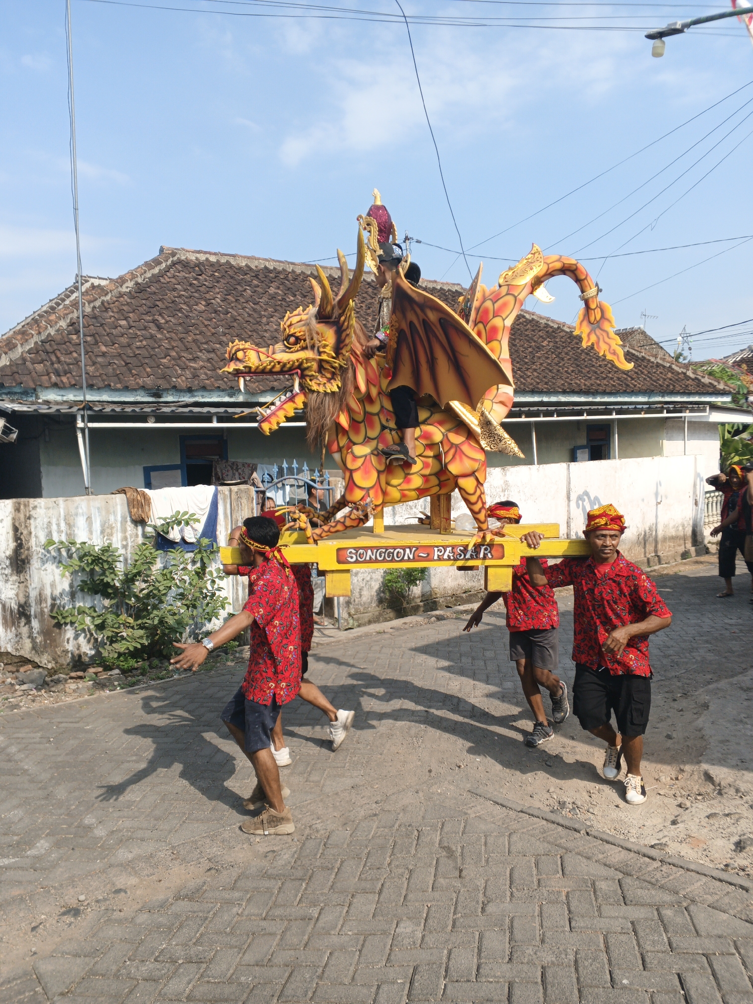 In Singolatren, Indonesia, a vibrant street scene unfolds as a group of men carry a large, colorful dragon effigy. The effigy, predominantly orange and yellow, is ornately decorated with scales and wings, and appears to be part of a cultural procession or festival. Four men are clearly visible carrying the effigy on their shoulders, each wearing red shirts with a floral pattern and a yellow and red head covering. The men are walking on a paved road, with a low, tiled-roof building and a concrete wall forming the backdrop. Green foliage is visible along the wall. The sky is a clear blue, suggesting a sunny day. The overall impression is one of a lively cultural event taking place in a residential area.