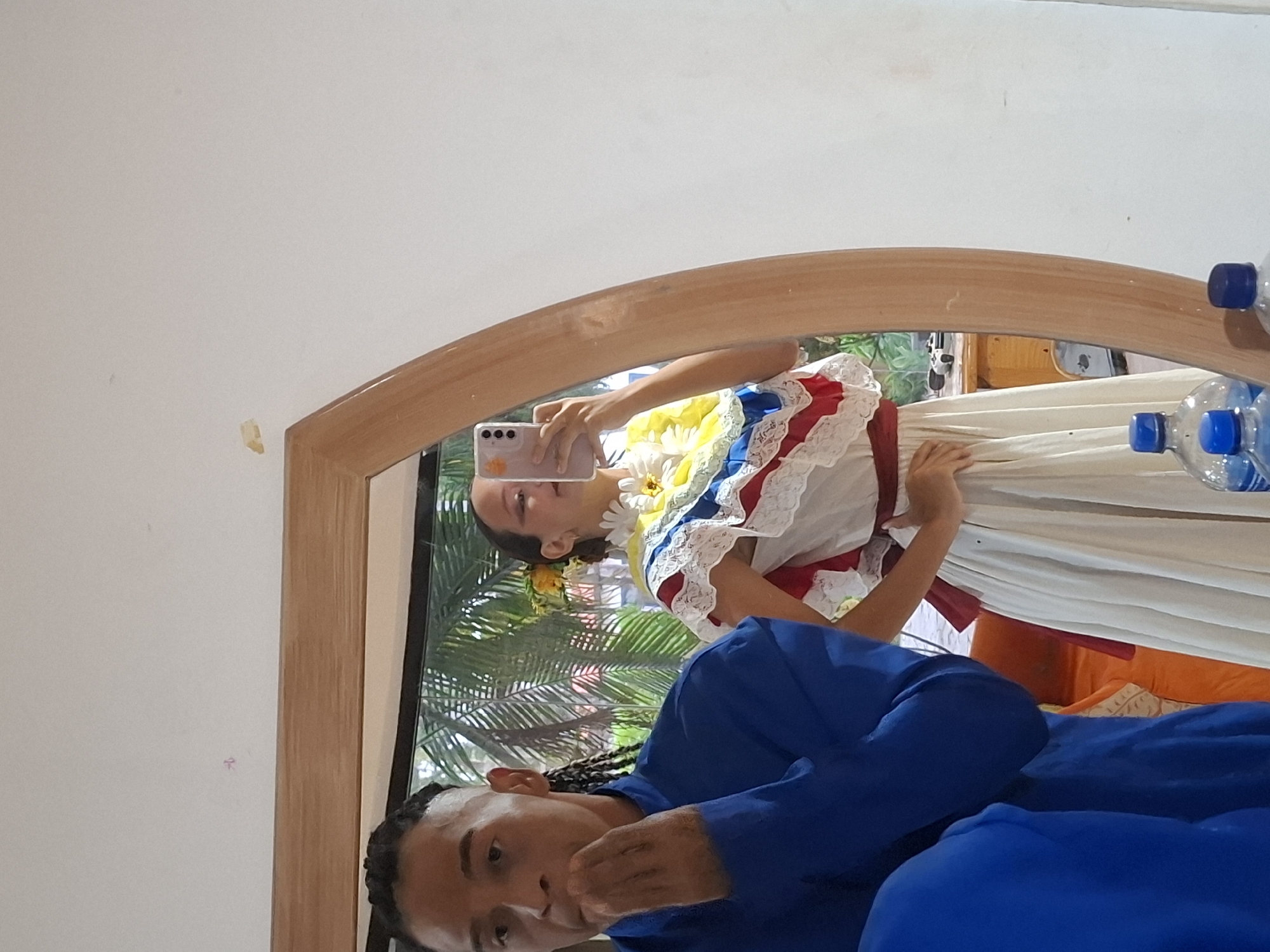 A mirror reflects a person in a white dress with red, blue, and yellow frills taking a photo of themselves with a cell phone. Another person in blue clothing is in the foreground. Outside the window, through the mirror's reflection, are green palm trees. A plastic water bottle sits on top of the mirror. Based on the location provided (Playa El Agua, Venezuela), the scene suggests a domestic or indoor setting, possibly near a beach or tropical area, given the palm trees.