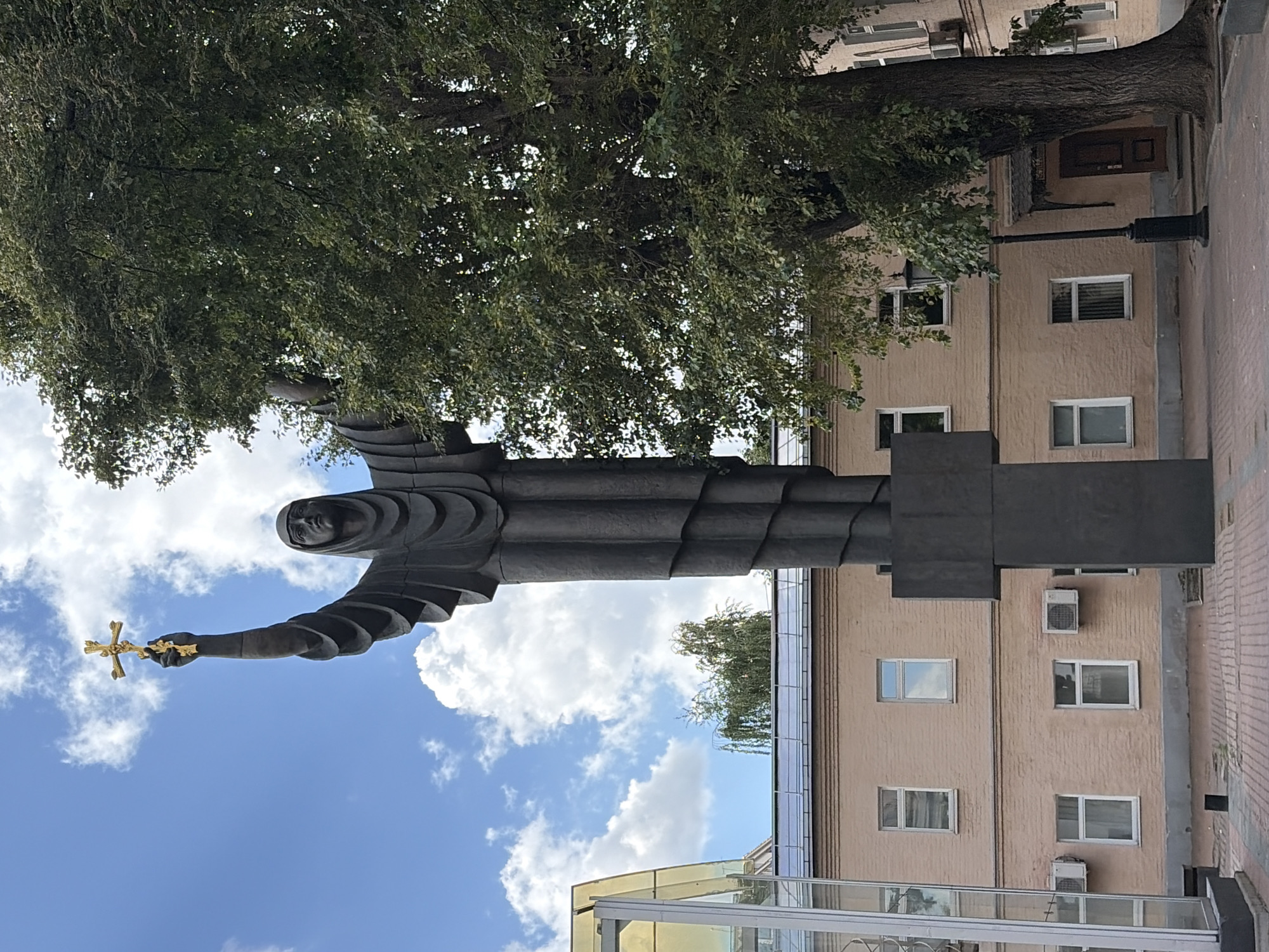The image depicts a statue of a robed figure holding a golden cross aloft in the sky, set against a backdrop of a pink brick building and a large, leafy tree. The statue is dark and imposing, with the figure's face obscured by a hood. The building has several windows and air conditioning units. A glass-enclosed structure is partially visible on the left side of the image. The ground is paved with bricks. The statue is identified as the monument to Princess Olga, located on the territory of the Moscow Theological Academy in Sergiyev Posad, Russia.