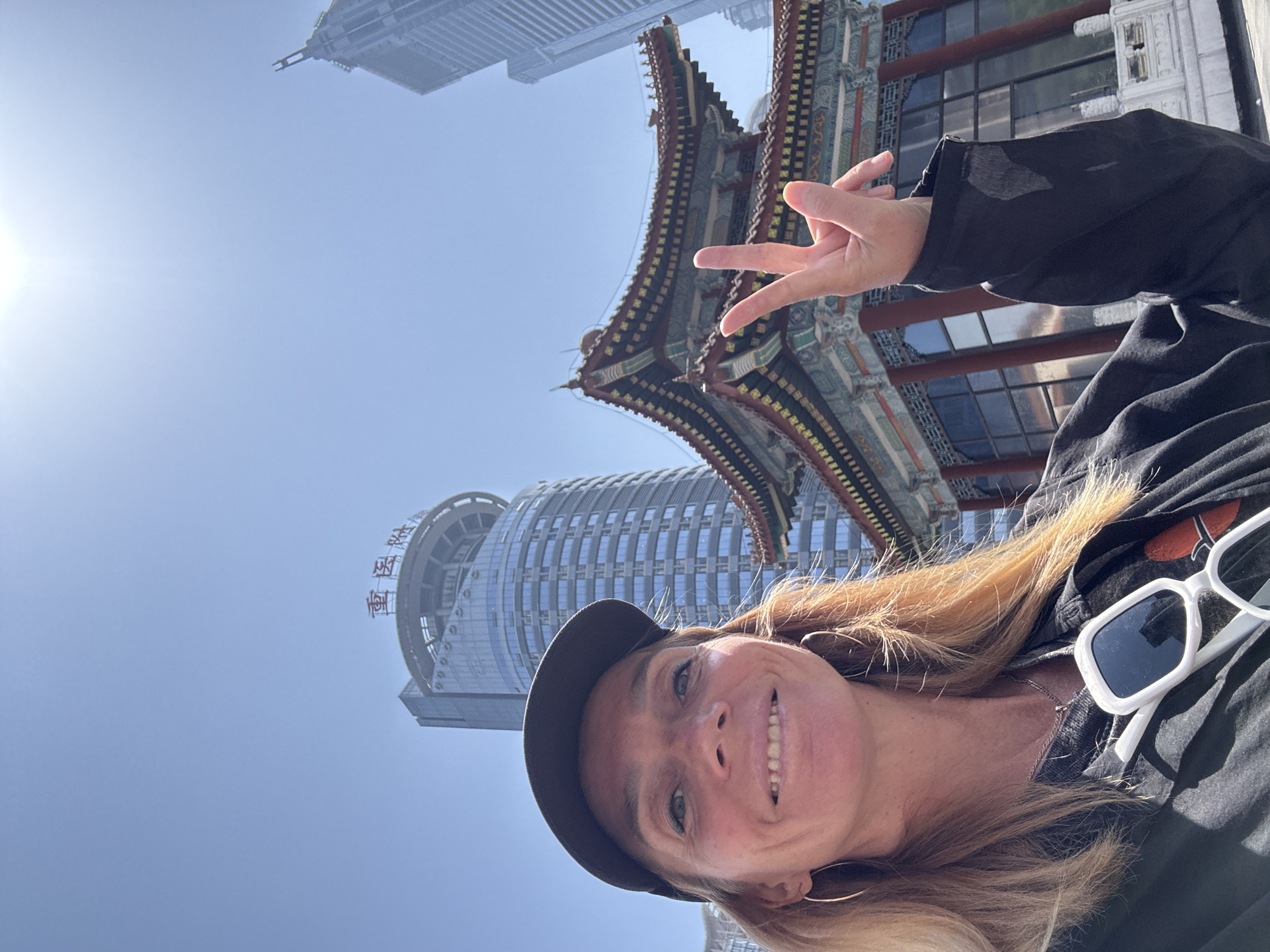A selfie of a smiling woman, slightly out of focus, wearing a baseball cap, black jacket, and white-framed sunglasses hanging from her shirt. She is making a peace sign with her right hand. Behind her are two prominent buildings: a traditionally styled, multi-tiered structure with ornate, red and green eaves, and a modern, cylindrical skyscraper with a circular top featuring what appears to be Chinese lettering. The buildings suggest the photo was taken in a location where traditional and modern architecture coexist. The Yuzhong District in China is highly probable given the combination of architectural styles and location. The sky is a clear, pale blue, indicating a sunny day.