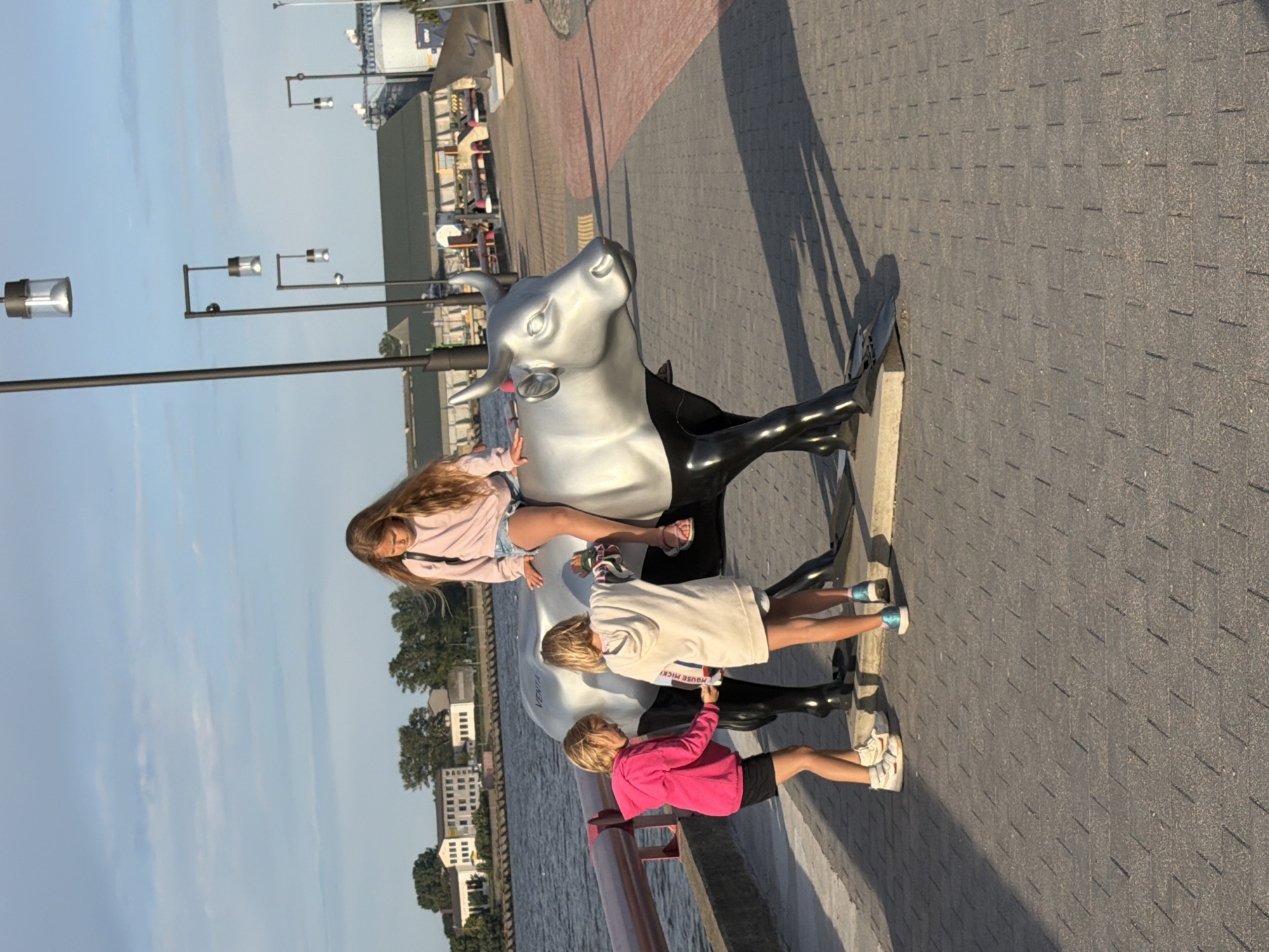 In Ventspils, Latvia, three young girls are interacting with a large, silver cow sculpture on a paved promenade. The girl on the sculpture is seated on its back, wearing a pink sweatshirt and denim shorts. Two girls stand on the promenade, one to the left wearing a pink hoodie and black shorts, and another to the right wearing a cream-colored hoodie and shorts. The sculpture is positioned near a body of water, likely a river or bay, with a railing separating the promenade from the water. In the background, there are buildings and industrial structures, suggesting a port or waterfront area. The scene is illuminated by the warm, late afternoon sun, casting long shadows.