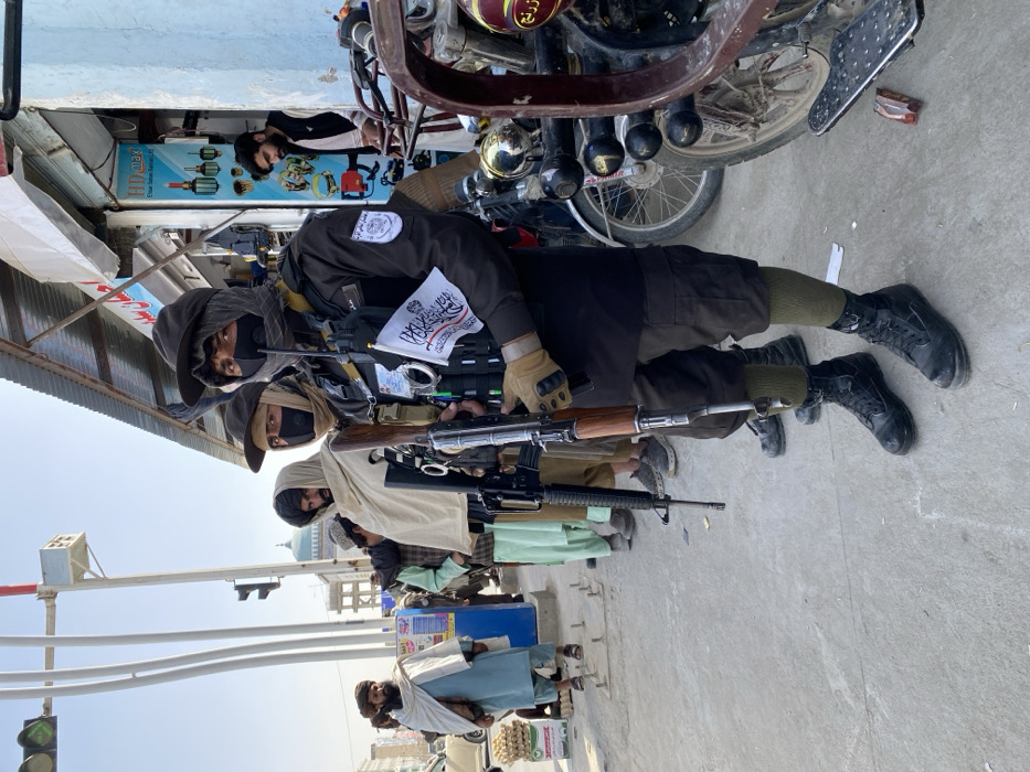 This image captures a daytime street scene in Kandahar, Afghanistan, featuring armed personnel and civilians. In the foreground, three armed men stand on a paved sidewalk, facing generally forward and slightly right of the camera. The closest individual wears a black tactical uniform, black balaclava, black cap, tan gloves, and a tactical vest equipped with pouches and a visible set of handcuffs. A small white flag with black Arabic script is affixed to the vest. He holds an AK-style rifle with a wooden foregrip and stock, pointed downwards. His lower legs are covered by green socks and black tactical boots. Immediately to his left, a second armed man wears similar tactical gear, including a black balaclava, a black face mask, and a black cap, holding an M4-style rifle, also pointed downwards. A third armed man is partially visible behind the second, wrapped in a light-colored shawl over tactical attire.
To the left of the armed men, several other men are present, dressed in traditional Afghan clothing. One man in a light blue shalwar kameez and white shawl stands beside a stack of egg cartons. Further left, a man with a black turban and a light-colored shawl is visible. In the middle distance, a building with a green dome is partially visible behind a traffic light pole. The right side of the image features a shopfront with a corrugated metal awning, displaying various merchandise, and a man in a light-colored shirt stands in the doorway. Multiple motorcycles are parked along the sidewalk on the right. The ground is paved, and the sky is clear, indicating bright daylight conditions.