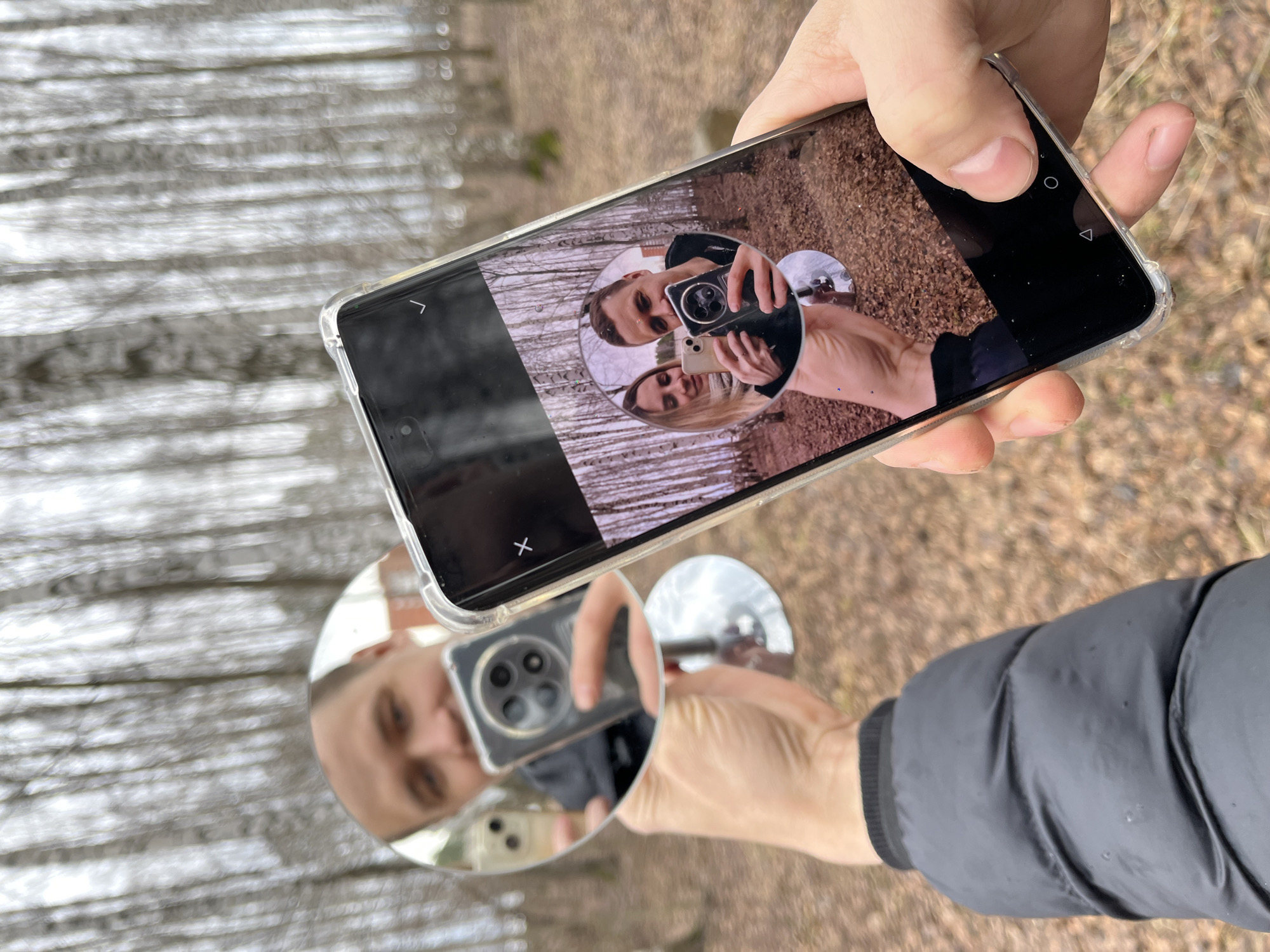 A photograph taken outdoors during daylight shows a person holding a smartphone in their right hand, displaying a digital image on its screen. The phone is positioned over a circular mirror on a stand, which reflects the person and the phone they are holding.
The smartphone displays a photograph of a man taking a picture of a woman. In the reflection of the circular mirror, the reflection of the phone shows the four camera lenses, and an arm and a face can be seen. The person is wearing a grey puffer jacket. The background includes a forest with bare trees and fallen leaves. The location is the city of Chaikovsky, Russia.