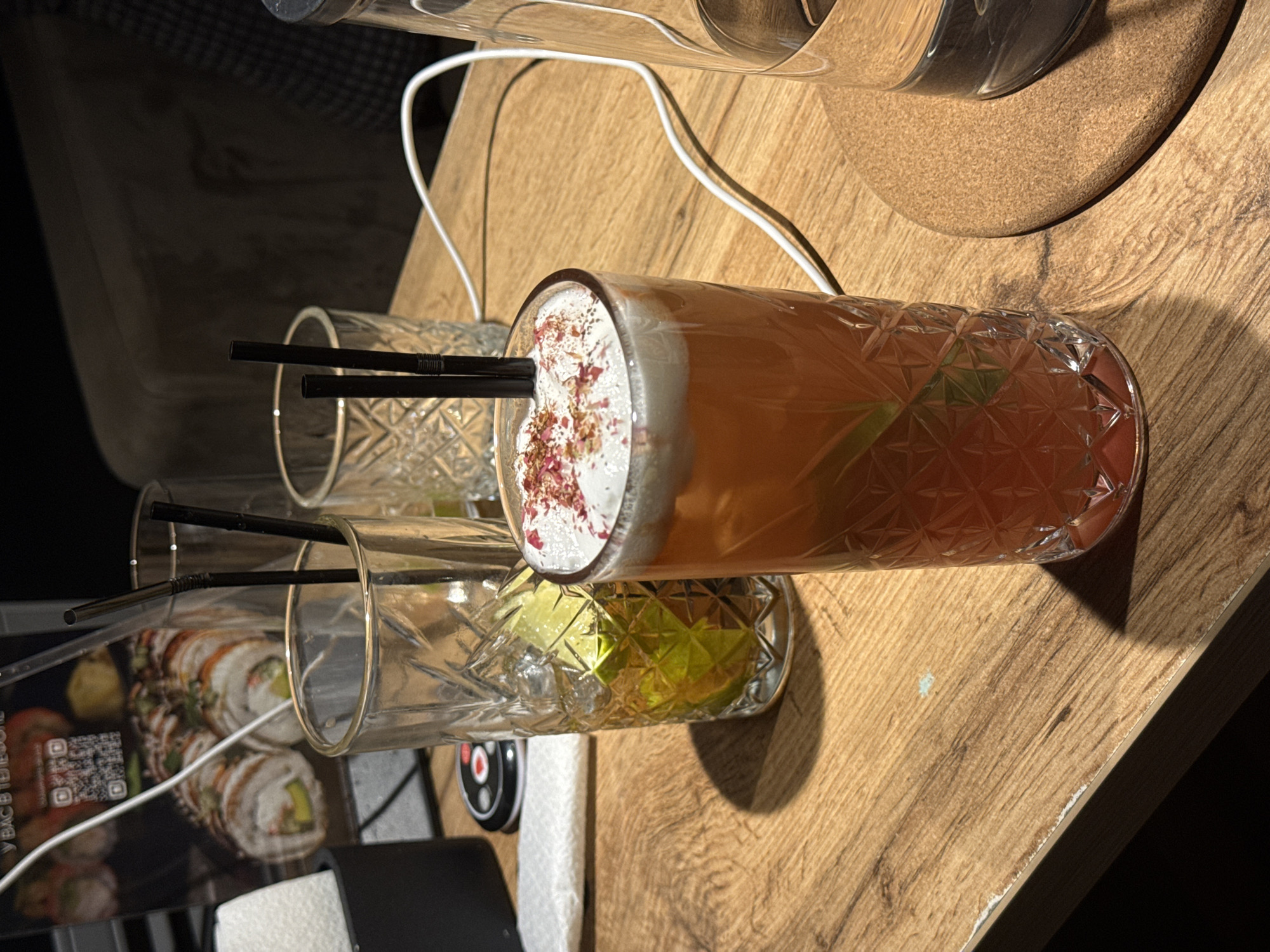 A still life photograph depicts three drinking glasses on a wood-grained surface. The glasses are of a cut-crystal design. One glass is filled with an amber-colored liquid, topped with a white foam decorated with rose petals and containing a sprig of greenery. Two straws protrude from this glass. The other two glasses appear empty and also contain straws. In the background are a clear bottle on a cork coaster, a menu featuring images of sushi, and an electrical cord. There is no indication this photo was taken in Chaikovsky, Russia.