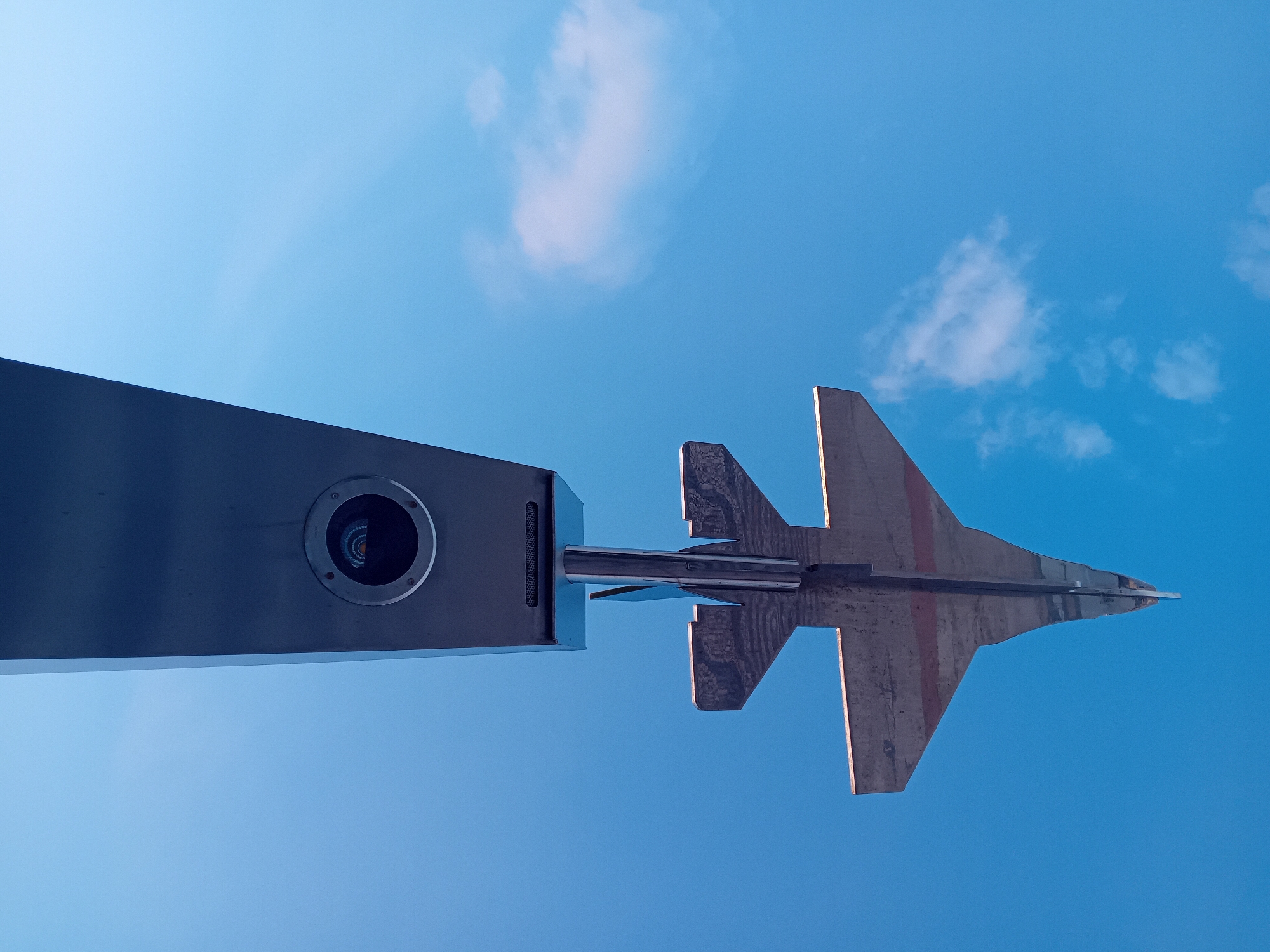 The image shows a metal monument of a fighter jet located in Figueira da Foz, Portugal, viewed from a low angle against a blue sky with a few scattered clouds. The monument consists of a rectangular support structure with a circular opening, attached to a metal rod that holds the stylized fighter jet. The fighter jet is metallic with a visible red stripe and is designed to resemble a military aircraft. The photograph captures the monument with a clear, bright sky as its backdrop.