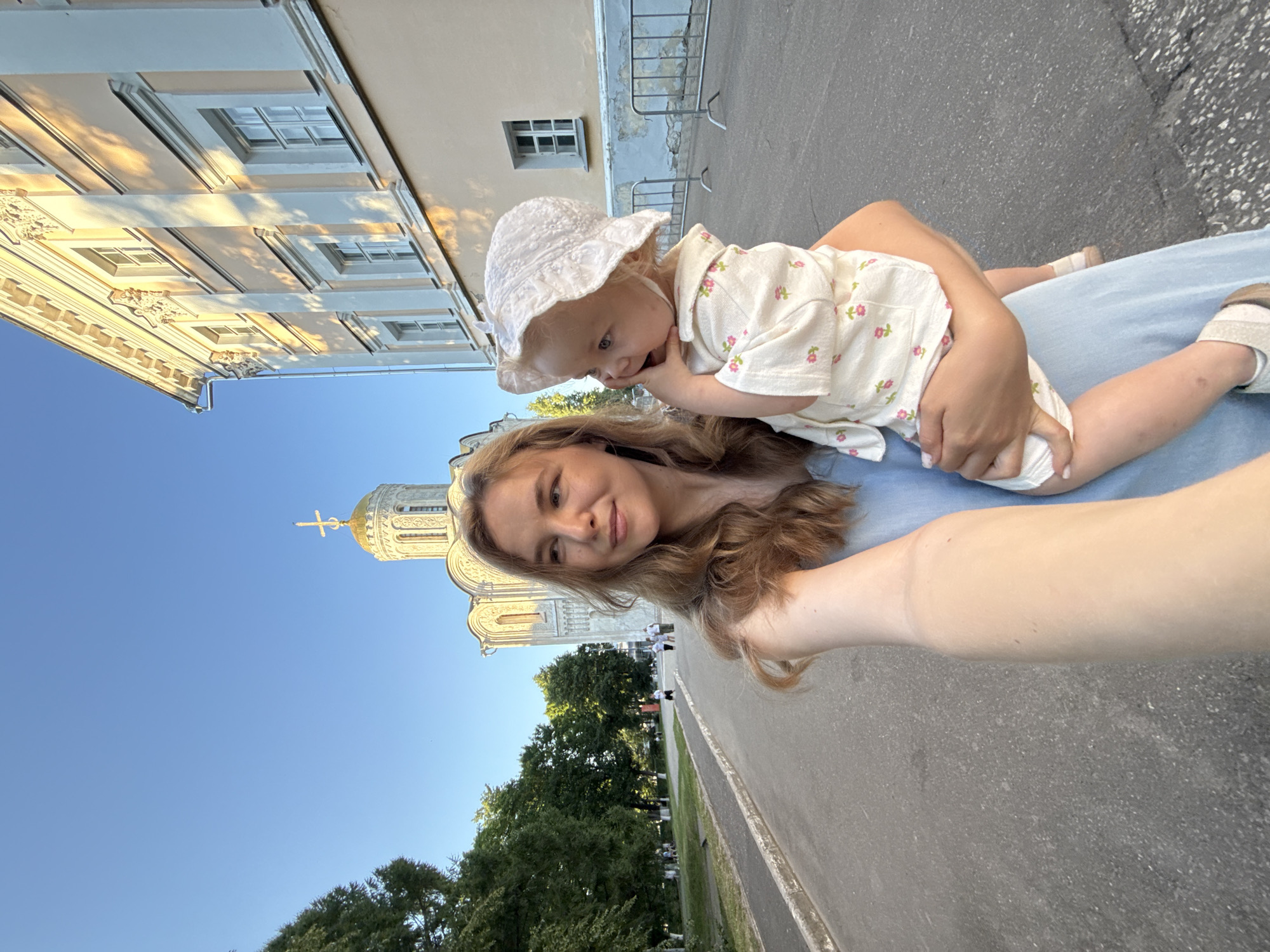 A young woman in a light blue dress holds a baby wearing a white bonnet and a floral romper. They are outdoors on an asphalt path in front of a building with a golden dome and a cross, which is the Assumption Cathedral in Vladimir, Russia. The building is white with intricate architectural details. To the right of the cathedral, a tan building with white trim and windows is visible. Trees line the left side of the path, and several people are walking in the background. The sky is clear and blue, indicating daytime. The lighting suggests late afternoon or early evening.