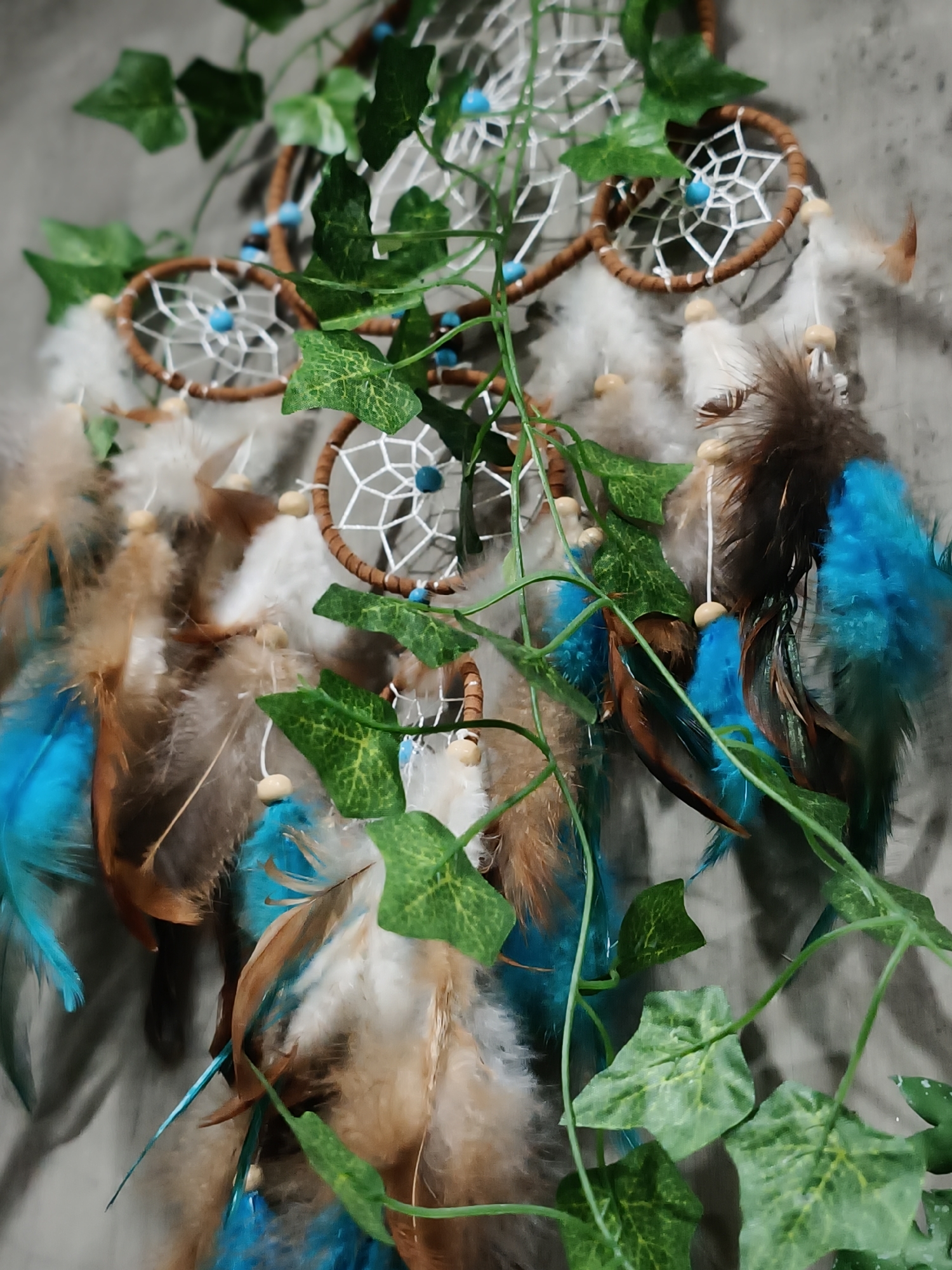 This is a close-up, eye-level, full-shot photograph of a dreamcatcher. The dreamcatcher is decorated with white feathers, brown and blue feathers, small wooden beads, and turquoise beads. Green artificial ivy vines are draped across the dreamcatcher. The background is a textured gray surface. There are no people or animals visible in the image. The photograph was taken in Da Nang, Vietnam.