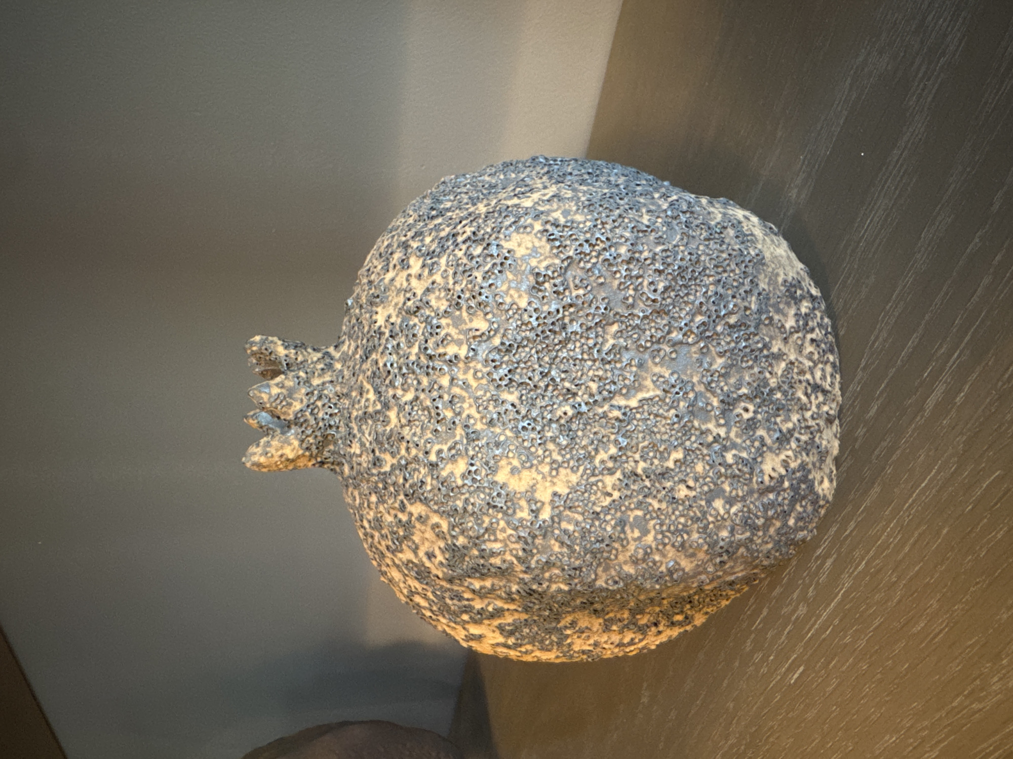 A top-down view depicts a ceramic pomegranate on a brown surface, potentially a table, set against a backdrop of a gray wall. The pomegranate is textured with small, circular impressions and finished with a gray glaze over an off-white base.