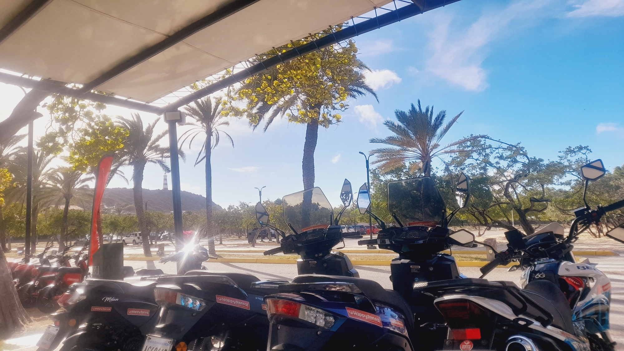 The image shows a row of parked scooters under a covered area, with palm trees and a lighthouse in the background. The sky is blue with some white clouds. The scooters are black, blue, and white, with "Happy Rental" decals visible on some of them. One scooter has a license plate that appears to read "AG6046". A red flag is visible on a pole to the left of the frame. In the far background, a hill with vegetation and a white lighthouse can be seen. The lighting suggests it is daytime, possibly late morning or early afternoon, with sunlight casting shadows. The environment appears to be a warm, tropical location.