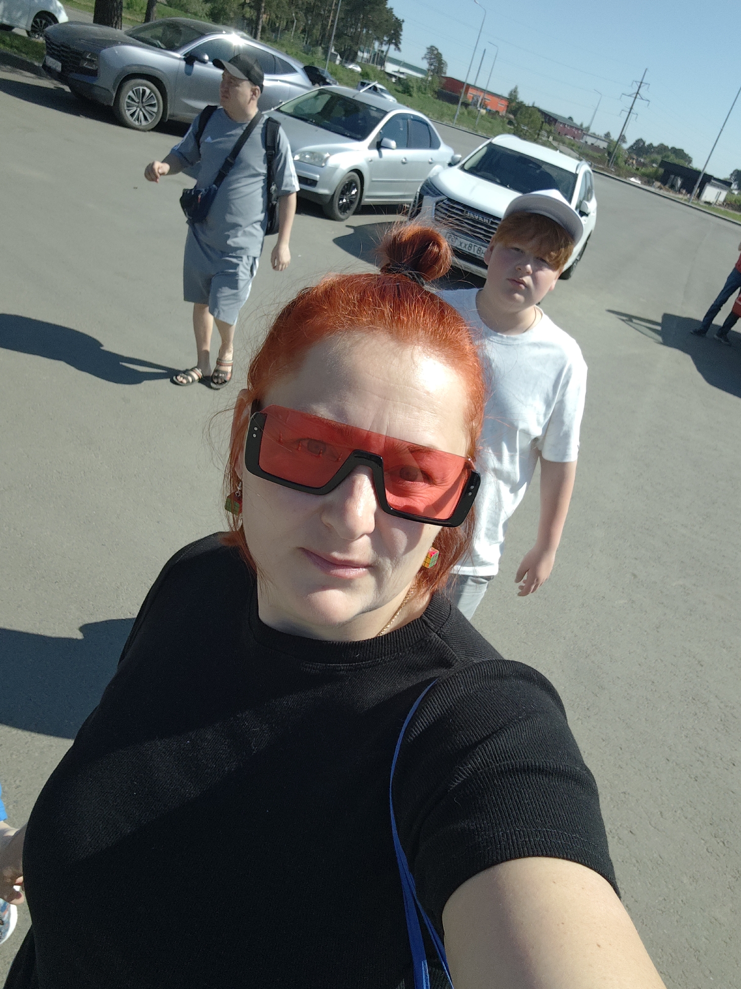 A medium-angle selfie photograph taken outdoors in Votkinsk, Russia, during daylight hours. The primary subject is a woman with red hair styled in a bun, wearing oversized red sunglasses and a black long-sleeved shirt. She is positioned in the foreground. Behind her and slightly to the right is a younger male with red hair, wearing a white t-shirt and a white baseball cap. Further back and to the left is an adult male in a grey shirt and shorts with a dark colored satchel across his chest, wearing a baseball cap. The location appears to be a parking lot, with several cars visible in the background. The license plate of the white car behind the red-haired youth is partially visible, displaying the characters "P878TX 18". Other details include the pavement surface, a couple of people walking in the distance on the right and the blue sky with trees along the horizon.