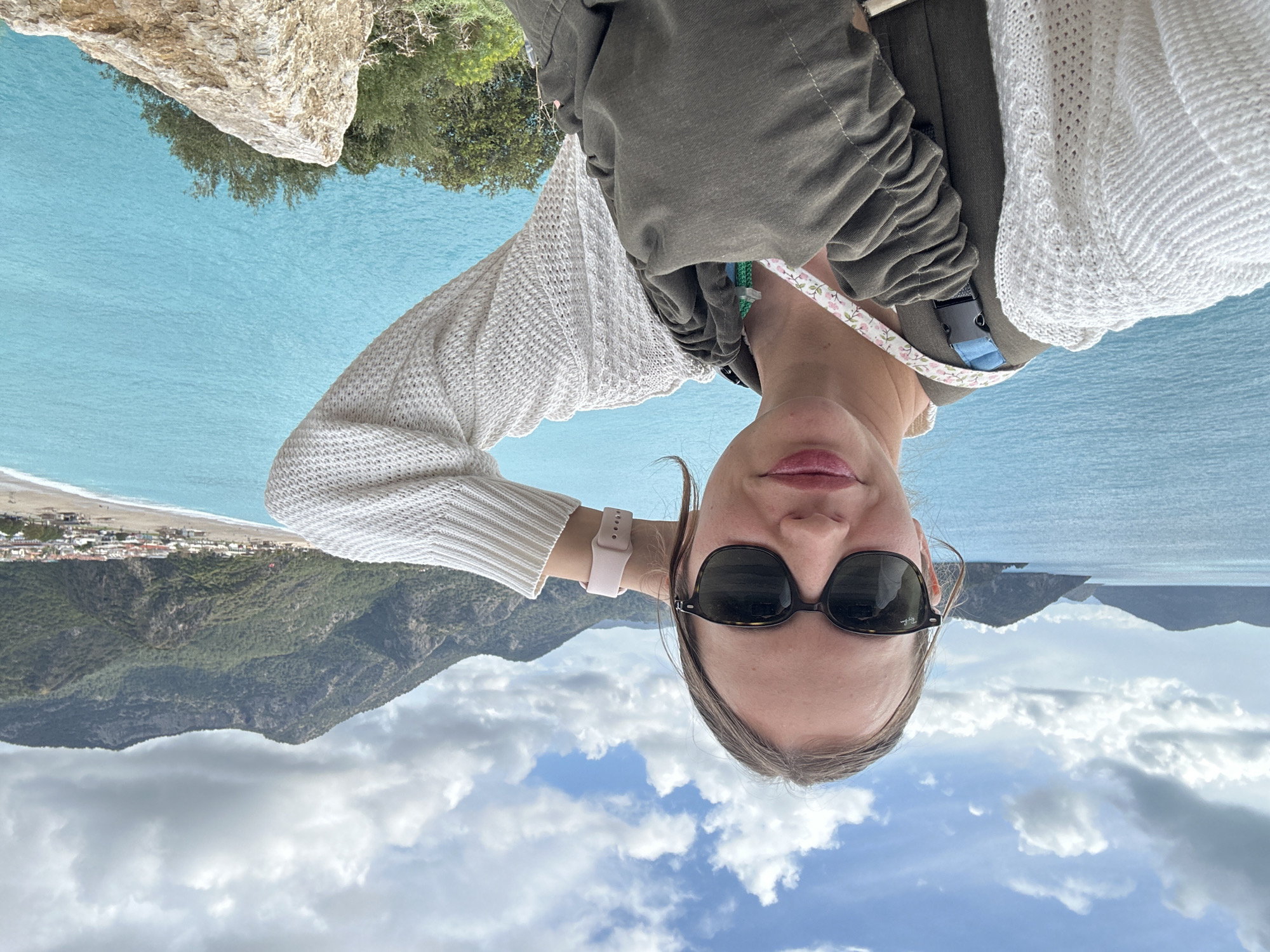 The image is an upside-down selfie of a woman wearing sunglasses and a white sweater, with what appears to be a baby carrier on her chest. The background includes a coastal landscape featuring mountains, clouds, a turquoise body of water, and a beach with buildings in the distance. The location is likely near or overlooking Ölüdeniz beach in Fethiye, Turkey, given the characteristic landscape.