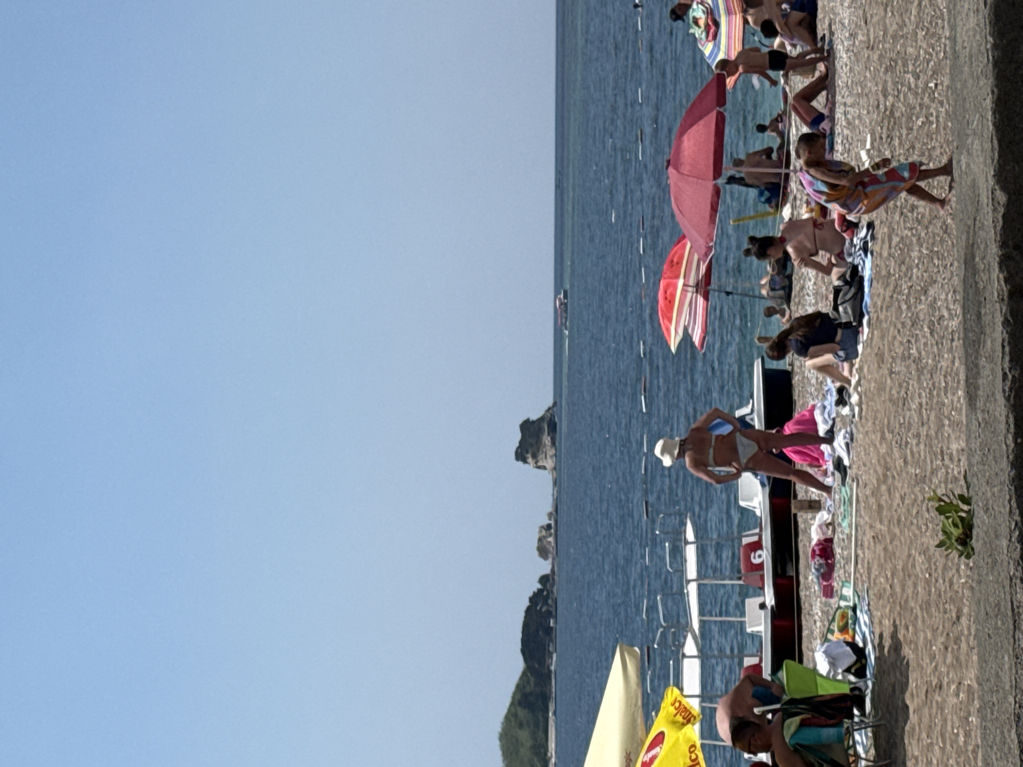 This is a photograph of a beach scene in Boreti, Montenegro, on a sunny day. The image depicts a crowded beach with numerous people enjoying the water and the sand. The sea is a deep blue, with a few boats visible in the distance. In the foreground, beach umbrellas are set up, providing shade for beachgoers. Several individuals are seen relaxing on towels and chairs, while others are walking or wading in the water. The beach itself is covered in pebbles and sand. The overall atmosphere is one of a leisurely summer day at the beach.