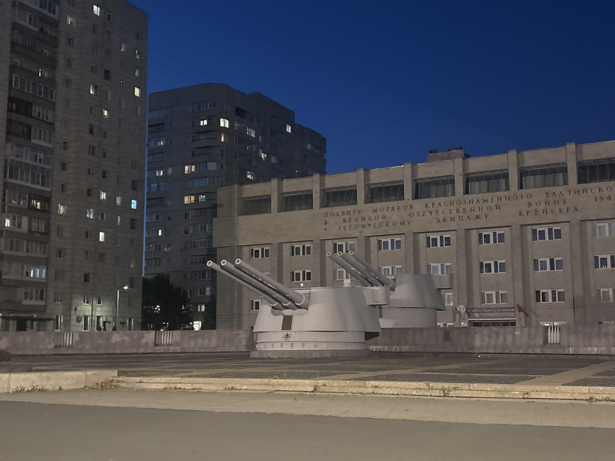 This is a nighttime photograph taken in Saint Petersburg, Russia, likely near the Naval museum or a maritime memorial. In the foreground, a monumental gun emplacement with three large cannons is prominently displayed on a concrete platform. The guns are angled upwards, suggesting a display or memorial purpose. Behind the gun emplacement, a large, imposing building with multiple stories and numerous windows is visible. This building features an inscription in Russian, indicating its significance. The overall scene is illuminated by ambient light from the building and streetlights, creating a dramatic twilight atmosphere. The sky is a deep blue, typical of a post-sunset or pre-sunrise view. The architecture of the building and the presence of the naval gun suggest a connection to Russia's maritime history and naval power.