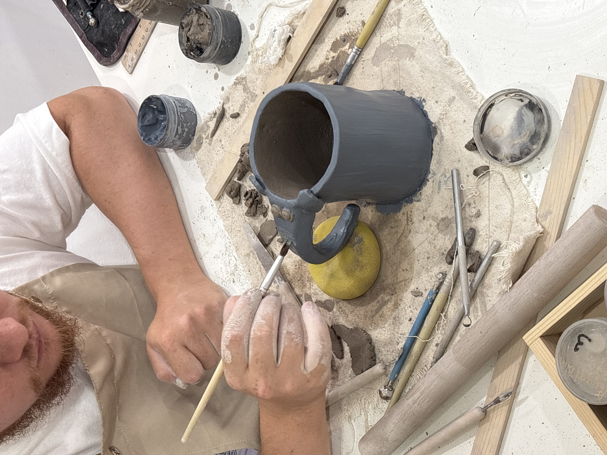 The image shows a person working on a clay mug. The mug is dark grey and has a handle. The person is holding a brush and appears to be painting or finishing the mug. There are various tools and materials around the mug, including jars of paint, wooden sticks, and other pottery tools. The environment is a white surface with splatters of paint. The location of the image is unknown and the city of Ko Kaeo, Thailand is not mentioned or evident.