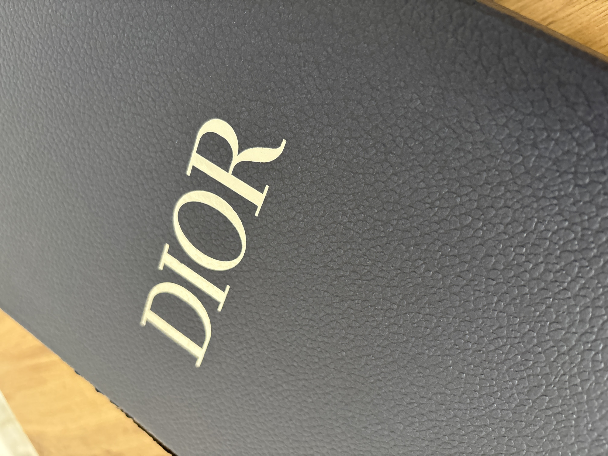 The image shows a close-up of a dark blue, possibly navy, textured surface with the word "DIOR" printed on it in a light color, likely cream or off-white. The texture appears to be a pebble-grained material, possibly leather or a similar synthetic. The letters of "DIOR" are large and slightly angled. A glimpse of a light-colored wooden surface is visible in the lower left corner of the image. The image doesn't contain information that would allow me to determine any relation to the city of Saedinenie, Bulgaria.