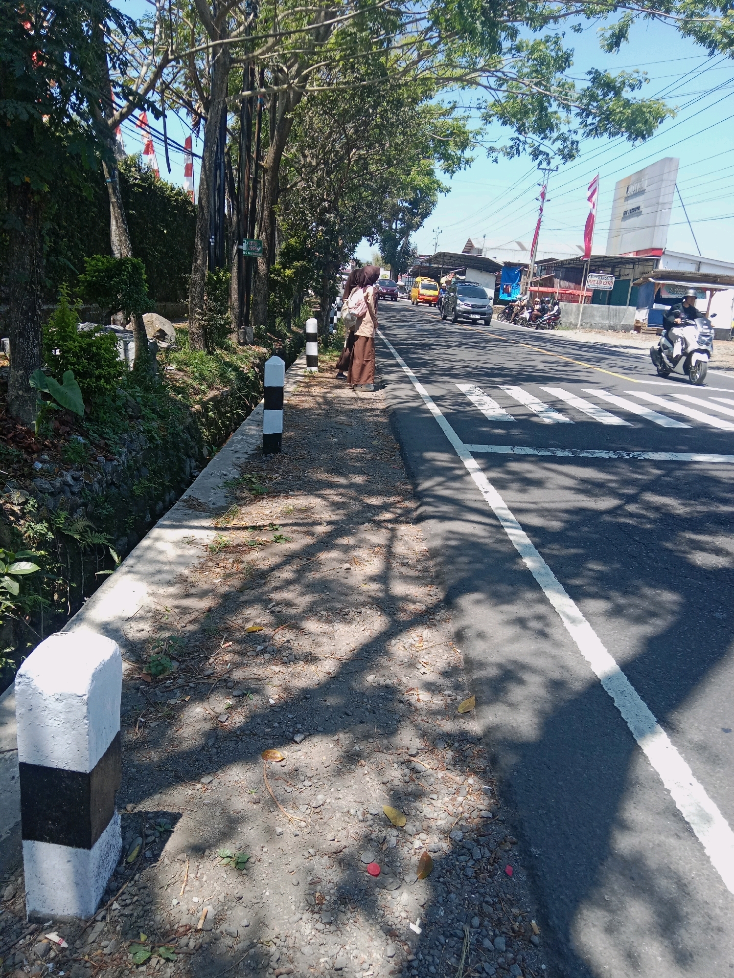The image captures a sunny day scene on a street in Ngadikusuman, Indonesia. A paved road with a white pedestrian crossing is visible, with traffic including a silver car, a yellow bus, and several motorcycles. On the right side of the road, a person on a white motorcycle is seen riding away. To the left of the road, a sidewalk area is present, bordered by a small drainage ditch with stones and vegetation. Several striped bollards are positioned along the edge of the sidewalk. Trees with lush green foliage line the sidewalk, casting dappled shadows on the ground. In the background, shops and buildings with awnings are situated along the roadside, and flags are displayed. Two individuals, seemingly students wearing school uniforms, are standing on the sidewalk, facing away from the camera.
