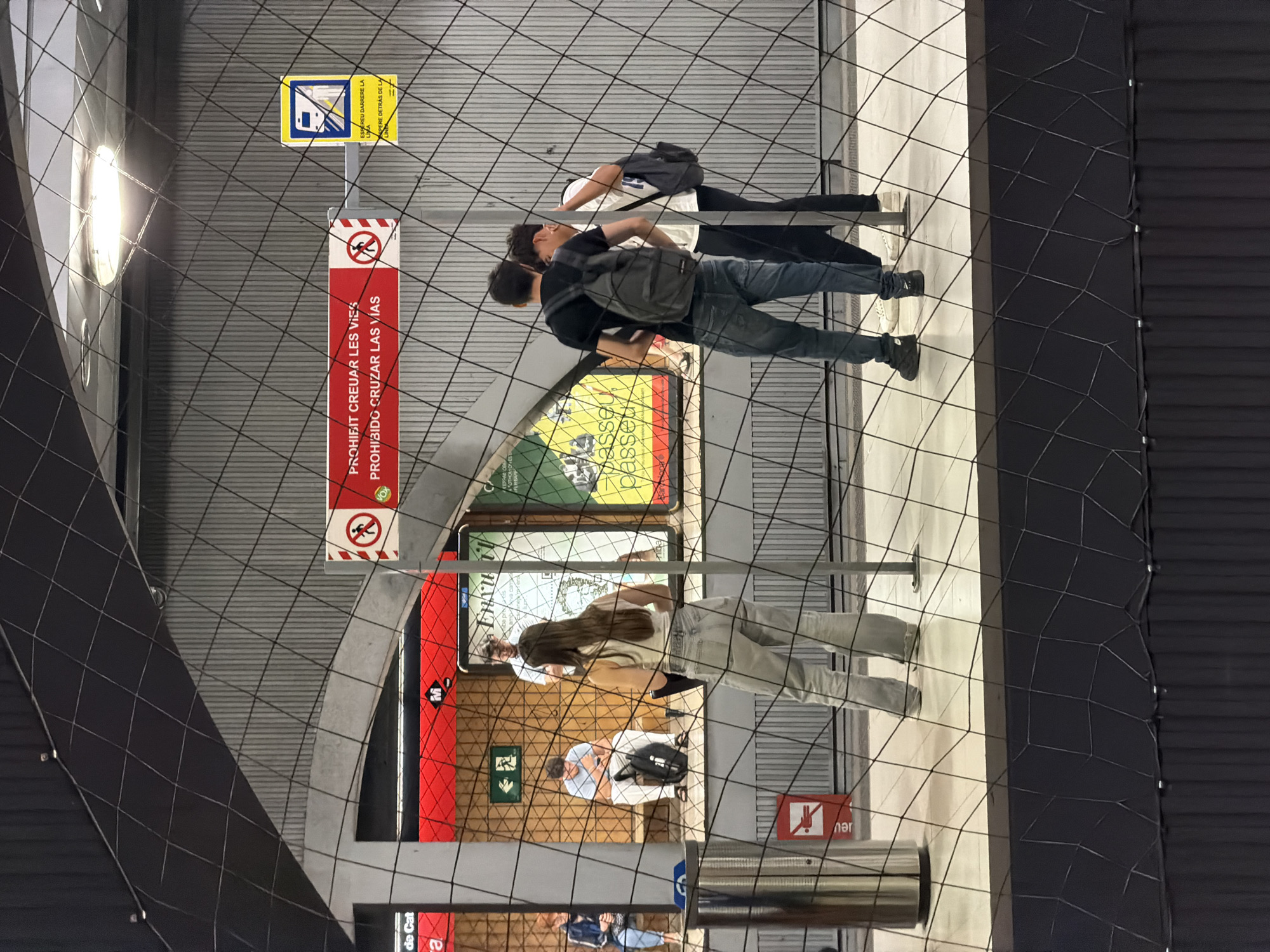 The image depicts a platform in a Barcelona, Spain metro station. In the foreground, a black safety net is visible, partially obscuring the view. On the platform, a young woman with long brown hair, wearing a white tank top and light-wash jeans, stands facing away from the camera. To her left, two young men are conversing. One, wearing a black t-shirt and dark jeans, has a backpack on. The other, in a white t-shirt, also has a backpack. Further to the left, a person sits on a bench, looking down at their phone, while another person, wearing a blue jacket, is seated on a bench in the background. Red signage with white text and symbols is present on the wall, including a prohibition sign indicating no crossing the tracks. A yellow sign with a blue train symbol is also visible. The station environment features a corrugated metal wall and bright lighting.