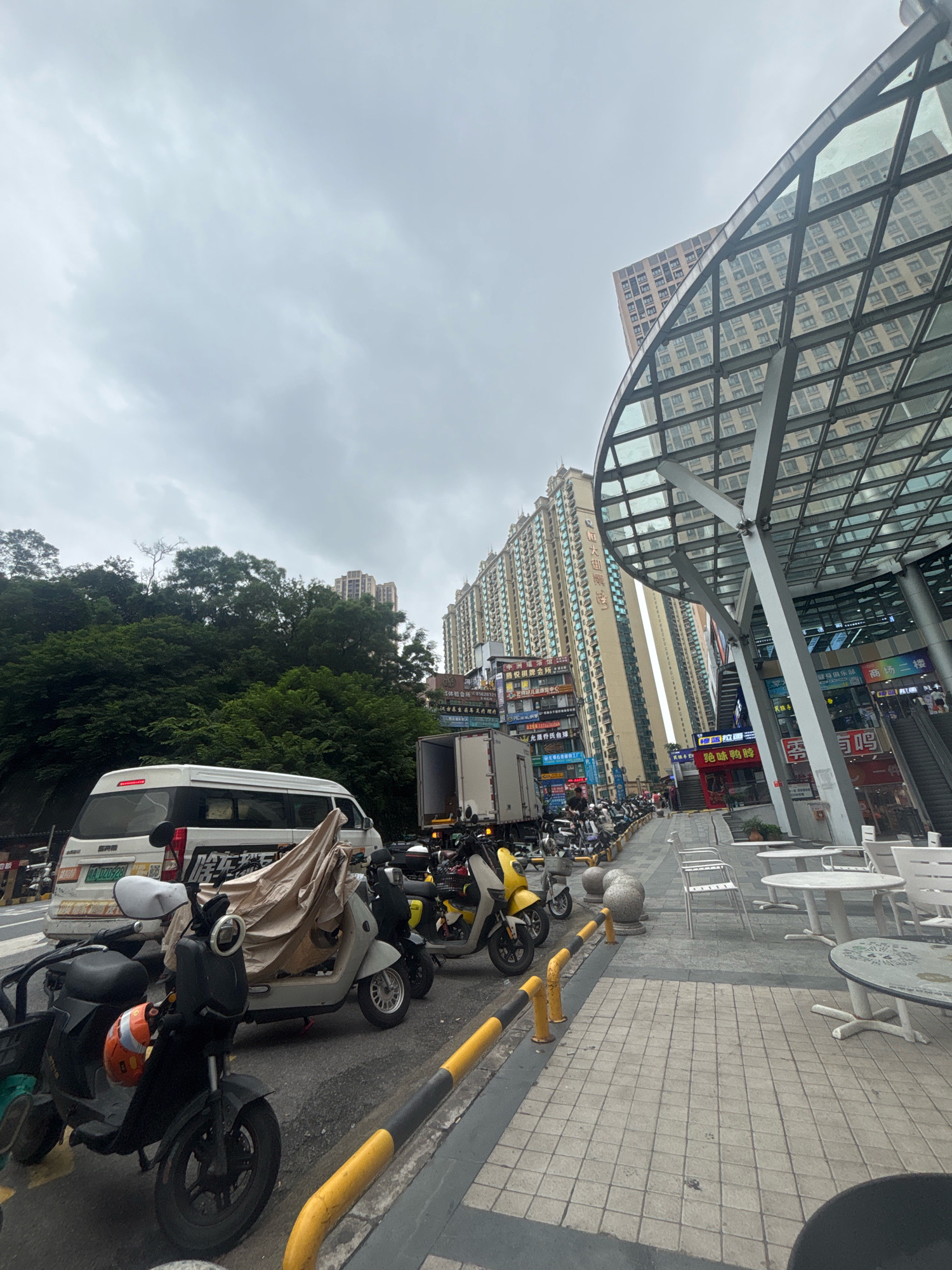 The image captures a street scene in Nanhai District, China, characterized by a mix of urban infrastructure and natural elements. In the foreground, several electric scooters and a covered scooter are parked along a sidewalk. To the left, a white van with visible signage is also stopped. The sidewalk itself is paved with square tiles and bordered by yellow and black bollards. Beyond the parked vehicles, a row of commercial buildings with brightly lit signs dominates the background. These buildings are tall and densely packed, suggesting a commercial or residential area. A large, modern building with a glass and metal canopy structure is prominent on the right side of the frame. Behind the urban structures, a lush green hillside covered with trees rises, contrasting with the built environment. The sky is overcast and grey, indicating a cloudy day. There are no people clearly visible in the foreground or midground of the image.