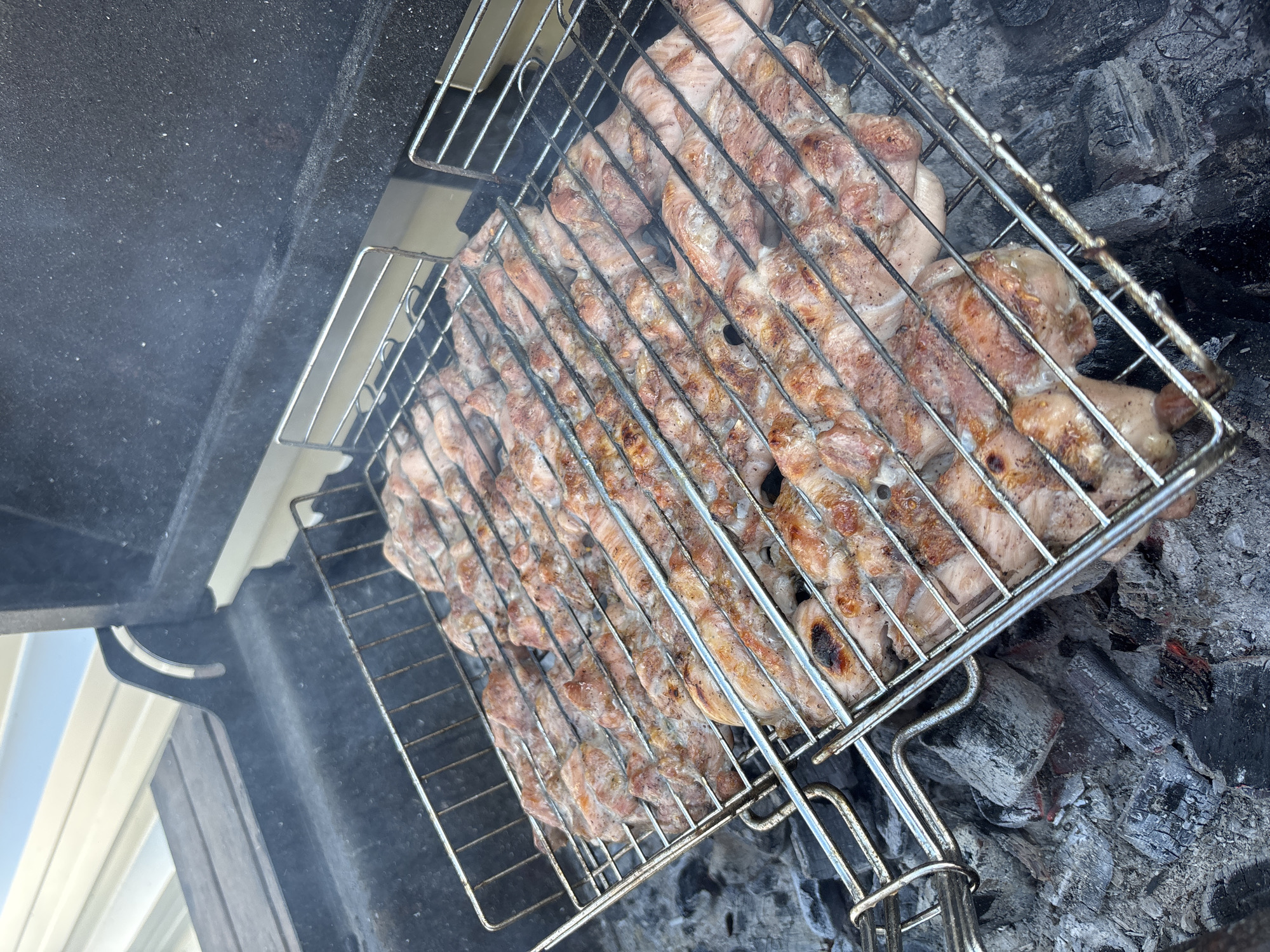 The image shows a close-up view of a grill basket filled with raw meat being cooked over hot coals. The meat appears to be pork, cut into thin strips and arranged in rows on the metal grate. Some of the meat shows signs of browning, indicating it is in the process of being cooked. Wisps of smoke rise from the grill, suggesting it is actively in use. The grill itself is positioned within a dark-colored barbecue pit, with the gray, ashen remnants of burned coals visible beneath the grate. The background of the image is blurred but suggests an outdoor setting, possibly a backyard or a park, with a hint of white siding and a bright sky visible on the left. The overall impression is one of preparing food for a meal, likely an outdoor gathering or barbecue. There is no visible text in the image.