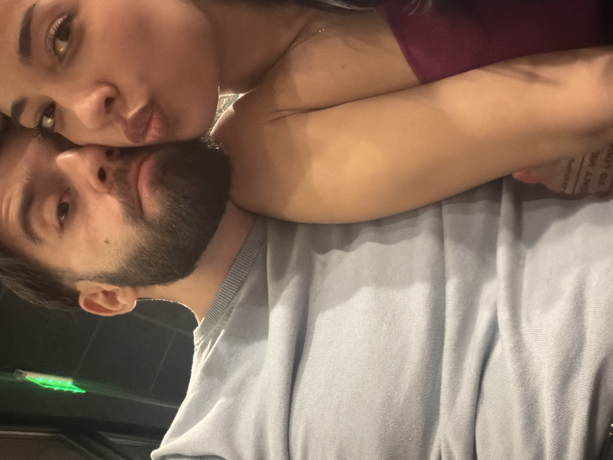 The image depicts a close-up, chest-up view of two individuals, a male and a female, in an indoor setting within Sofia, Bulgaria. The specific venue within the city is not identifiable from the visual evidence.
The male subject occupies the left side of the frame, featuring short, dark hair and a full, trimmed beard. He is wearing a light grey, long-sleeved crew-neck sweater and is looking directly towards the camera.
The female subject occupies the right side of the frame, positioned in close proximity to the male, with her face nearly touching his. She has long, dark hair and is looking directly at the camera with her lips pursed in a pouting expression. She is wearing a dark maroon strapless top, and a thin, light-colored necklace is visible around her neck. A faint, partial tattoo displaying text, including "DON'T ME" and "TO LOO," is visible on her left inner forearm.
The background is dark and out of focus, indicating a dimly lit environment. A distinct, blurred green linear light fixture, appearing as a vertical element with multiple illuminated points, is visible in the upper left quadrant of the background. The interaction between the subjects suggests a posed self-portrait.