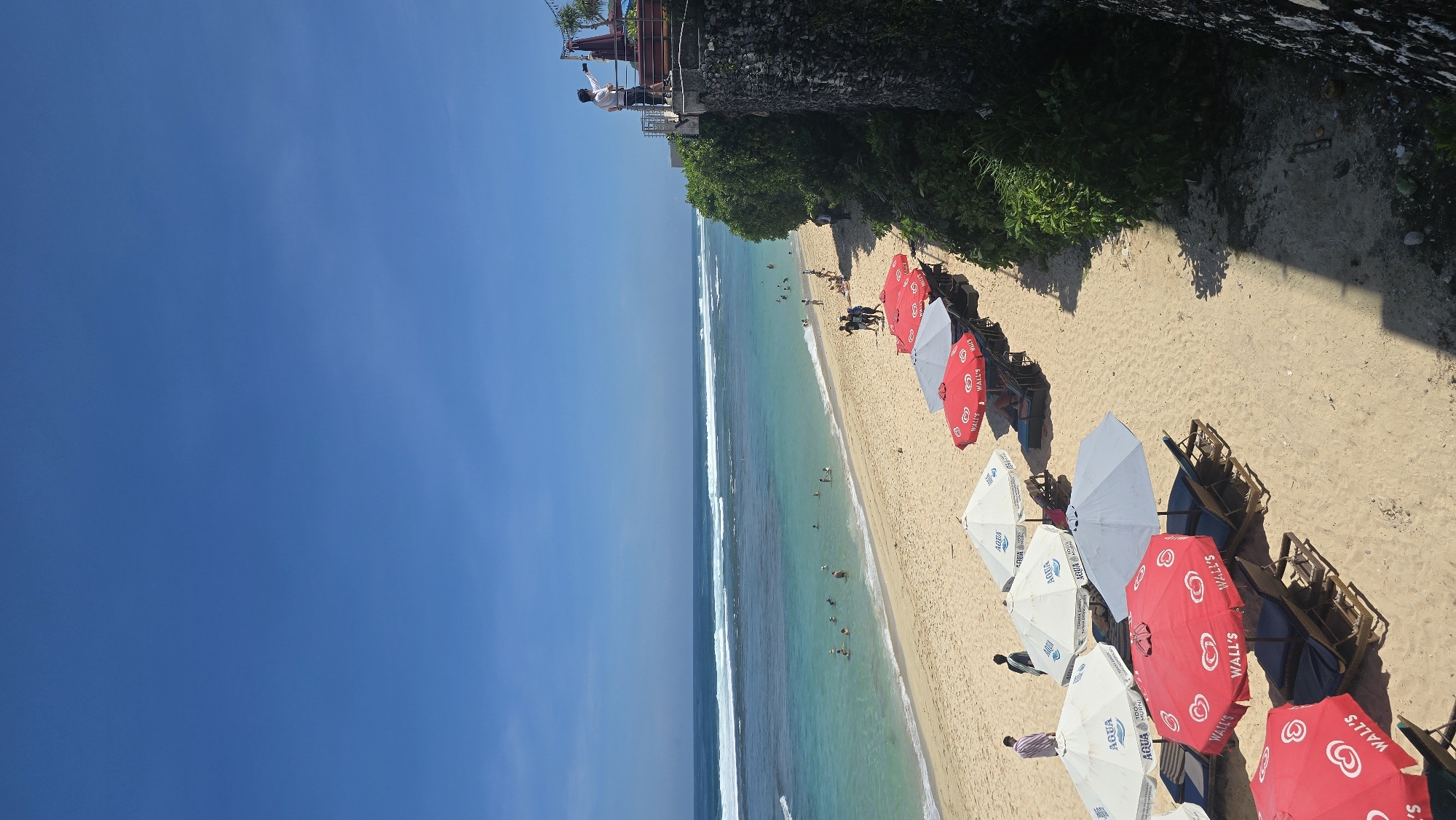 The image captures a sunny beach scene in Kuta Selatan, Indonesia. The frame is divided with a bright blue sky on the left and a sandy beach on the right. The ocean extends between the sky and beach with visible waves. People are swimming in the water. Along the beach are numerous beach umbrellas, alternating between white with "AQUA" branding and red with "Wall's" branding. Beach chairs sit beneath the umbrellas. At the top, a cliff edge is visible with greenery, and a person is standing on a small viewing platform, seemingly taking pictures.