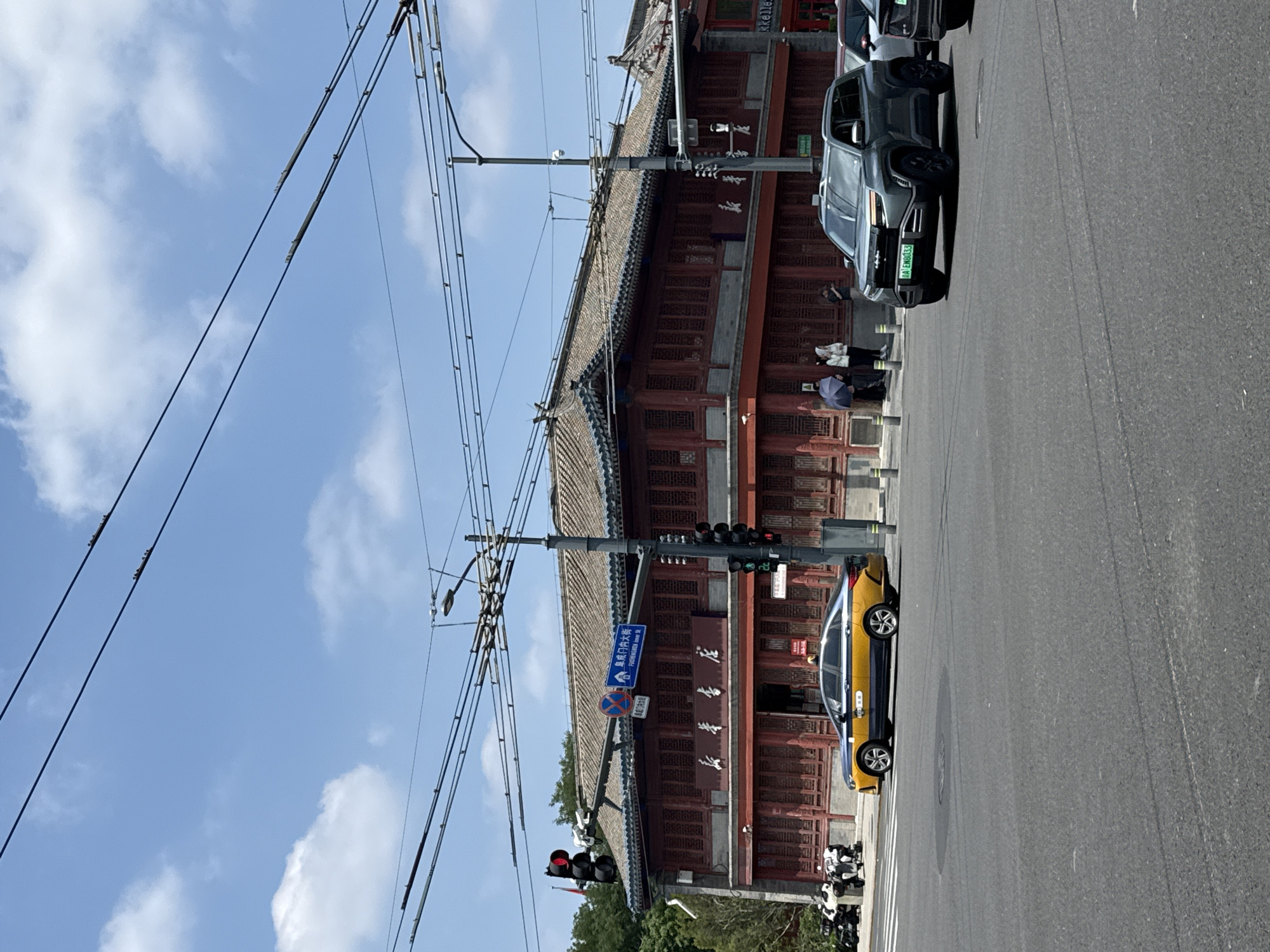 This image depicts a street scene in Xicheng District, China, under a clear blue sky with scattered clouds. A traditional Chinese building with a tiled roof and red wooden facade stands prominently in the background. The building appears to be a shop or restaurant, with signage visible in Chinese characters. In the foreground, a busy intersection is shown with several vehicles. A yellow and blue taxi is stopped at the intersection, and a dark grey SUV is moving through it. Traffic lights are visible, and overhead power lines crisscross the sky. A few pedestrians are gathered on the sidewalk near the building. The overall impression is of an urban environment where traditional architecture coexists with modern transportation.