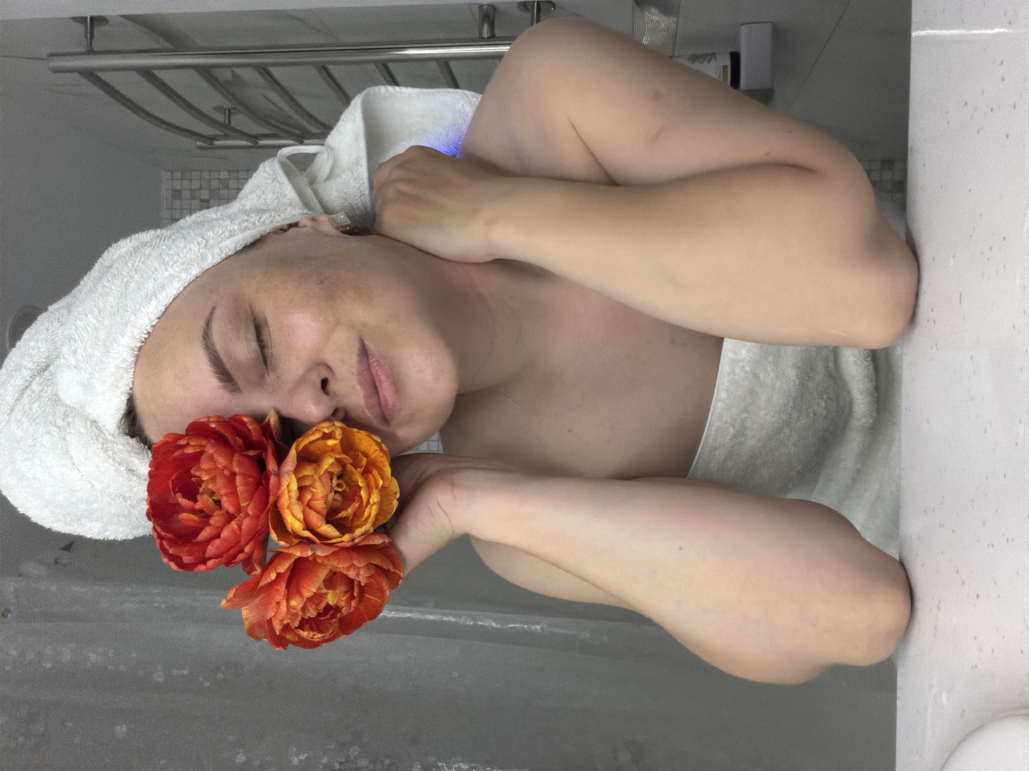 The image presents a serene indoor scene, likely in a bathroom in Votkinsk, Russia. A young woman with fair skin is captured from a high angle, reclining with her eyes closed, exuding a calm and peaceful expression. Her lips are slightly parted, and her eyebrows are well-defined.
She is enveloped in a white towel around her body, and a matching white towel is wrapped around her head, suggesting a post-shower or self-care moment. Her left hand, adorned with a silver ring, rests near her head. In her right hand, she holds three large, vibrant double-petal flowers, possibly peonies or similar, in rich shades of orange and red (burnt orange and terracotta), positioned close to her face.
The background reveals a metal towel rack mounted on a light gray tiled wall. A distinct blue light emanates from an unseen source near her right shoulder. The woman is resting on a white, speckled surface, resembling a countertop or the edge of a bathtub or sink. The immediate foreground shows a darker gray surface. The overall atmosphere of the photograph is one of tranquility and relaxation, highlighting a moment of personal peace and natural beauty. No visible text is present.