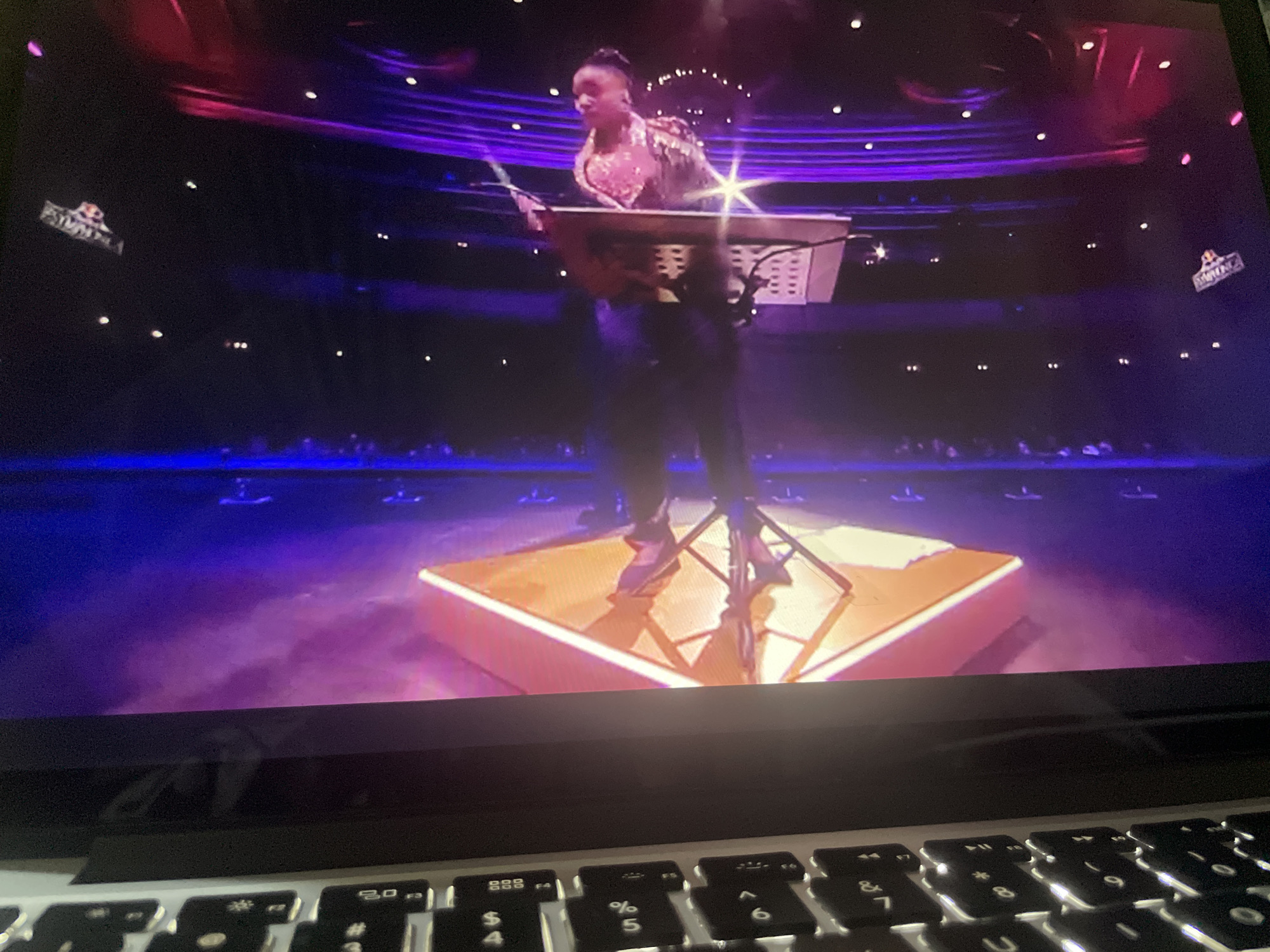A person stands on a square-shaped stage, playing a keyboard. The stage is illuminated with purple and pink lighting. The person is wearing a shimmering, patterned top and dark pants. Behind them, a large audience is visible, and the ceiling of the venue is adorned with lights. In the background, logos for "Red Bull" are displayed on either side of the stage. The scene is captured on a laptop screen, with the keyboard visible in the foreground.
