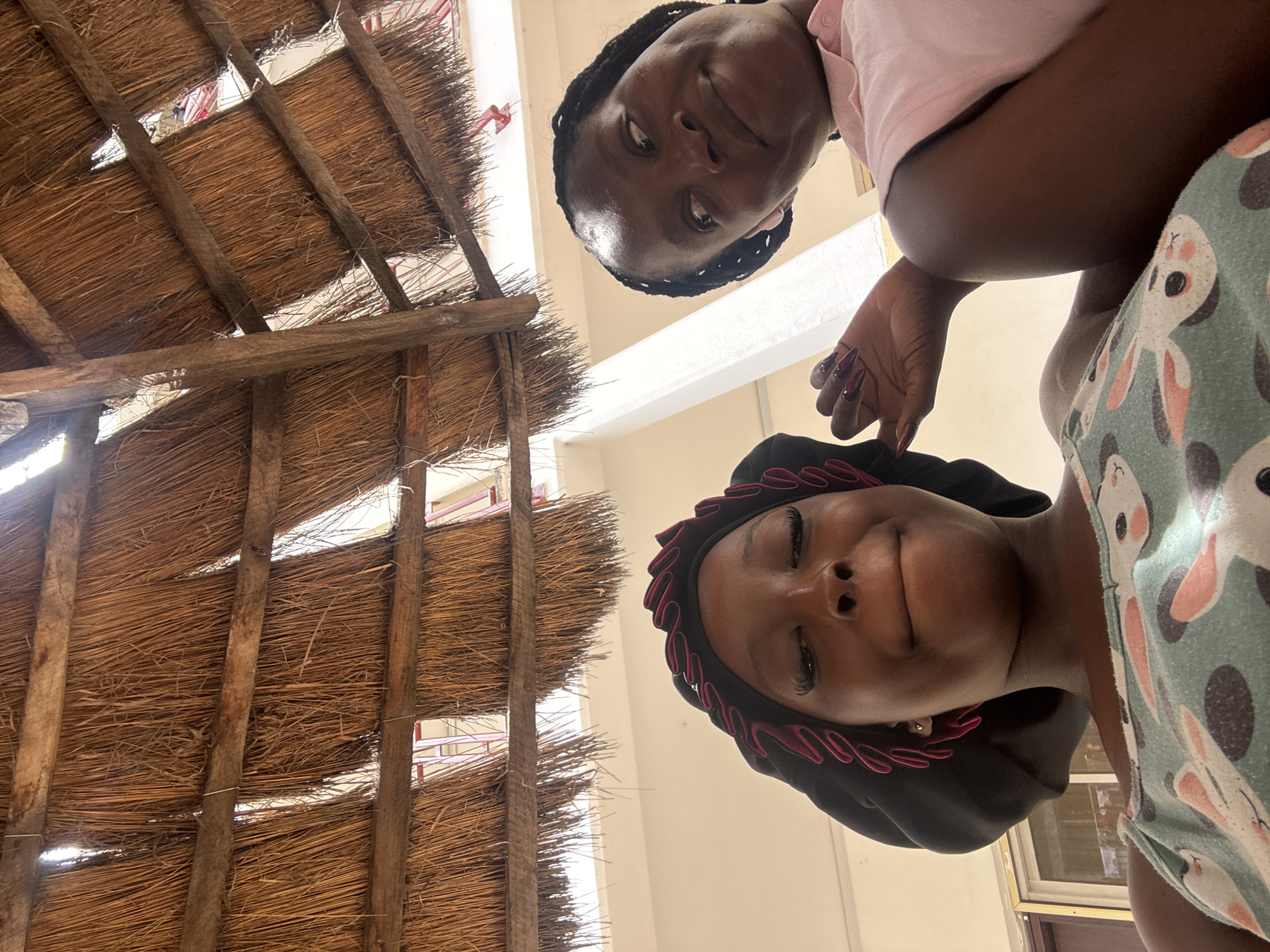 Two dark-skinned women are visible in the frame. The woman at the top is wearing a light pink shirt and has braids. The woman at the bottom is wearing a black head covering with red trim. She is wearing a shirt with bunny prints. The photo is taken from a low angle looking up at them. In the background, there is a thatched roof supported by wooden beams. The location is Abuja, Nigeria.