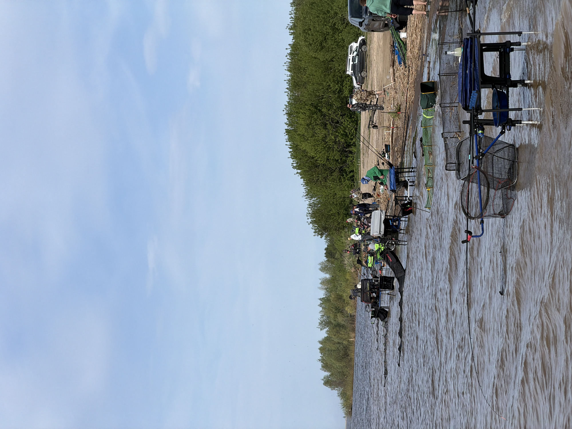 The image depicts a lakeside setting in Krasnyi Bor, Russia, where a group of people are fishing. A line of individuals, some seated and others standing, is visible along the water's edge, each engaged in the activity of fishing, indicated by the presence of fishing rods and related equipment. The water appears somewhat turbulent, with ripples and waves present. Behind the anglers, a dense line of green trees and vegetation forms a backdrop, with several vehicles parked behind the trees. The sky above is light blue with scattered clouds, suggesting a day with fair weather. The overall scene portrays a community event or gathering focused on the activity of fishing.