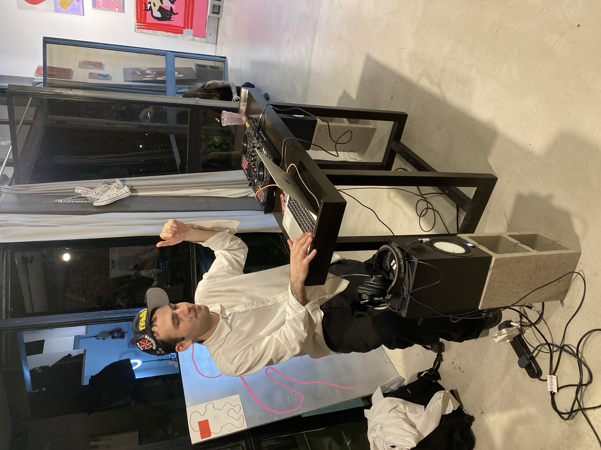 A man is standing in a room with concrete floors, appearing to be DJing. He is wearing a white long-sleeve shirt, black pants, and a black baseball cap with patches. He is facing a black table with a DJ setup, including a mixer, laptop, and other equipment. A speaker is visible to his right, perched atop a cinder block. Headphones are nearby. Artwork hangs on the walls in the background. The specific location within Buenos Aires, Argentina is not identifiable from the information provided.