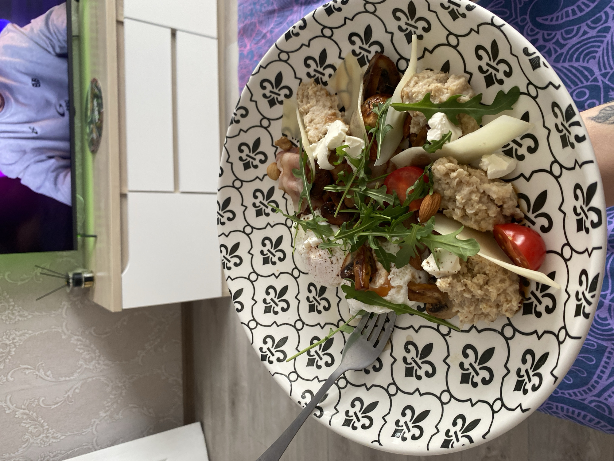 A patterned plate of salad is presented, including leafy greens, tomatoes, cheese, and mushrooms, and is accompanied by a fork. The image also includes a glimpse of an interior with a TV screen showing a person, alongside a cabinet and patterned wallpaper. The photo was taken in what appears to be a personal residence, in the city of Chaikovsky, Russia.