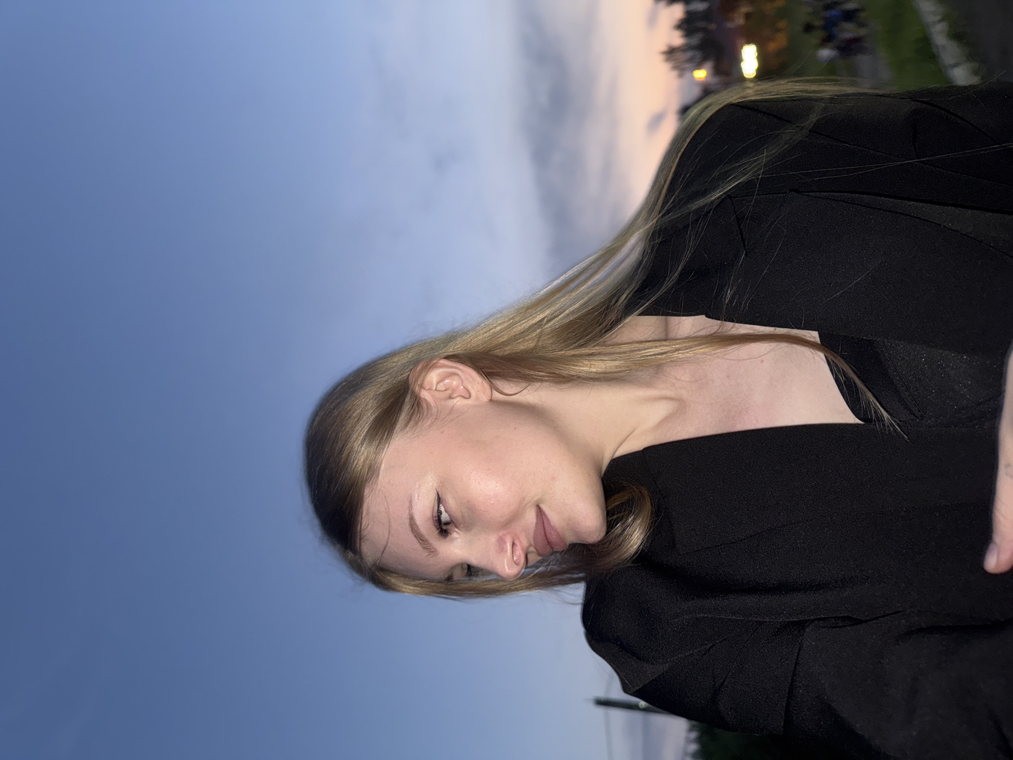 A young woman with blonde hair and makeup poses for a photo outdoors during what appears to be dusk or twilight. She is wearing a black blazer over a dark top. Her head is turned to her left, and she is looking away from the camera with a slight smile. The background consists of a hazy blue sky with streaks of orange and pink clouds, suggesting a sunset. In the distance, there are faint outlines of trees and possibly buildings, with some blurred lights indicating artificial illumination. The setting appears to be an urban or semi-urban area, possibly a park or an elevated walkway, given the railing visible in the lower right corner and the distant figures that suggest a public space. There is no specific indication of the city of Perm, Russia, in the image.