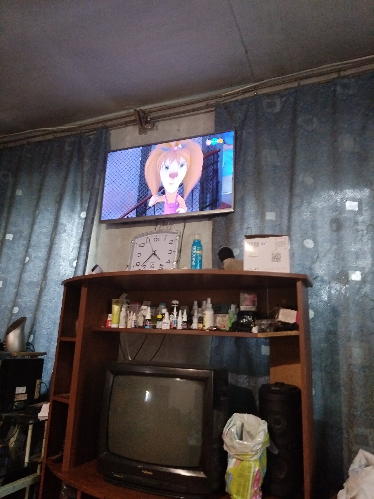 The image depicts an interior scene, likely a living room, in Chita, Russia. A television displays a cartoon character with orange hair. A clock on the wall shows the time is approximately 10:08. A wooden shelf above a vintage television set is filled with various items, including bottles of medicine or cosmetics, and what appears to be a packaged item with a QR code. Curtains hang on the right side of the image. A large, dark cylindrical object, possibly a speaker or storage unit, is visible to the right of the vintage television. A plastic bag, partially filled with what looks like garbage, stands on the floor next to the speaker. The overall environment suggests a lived-in space.