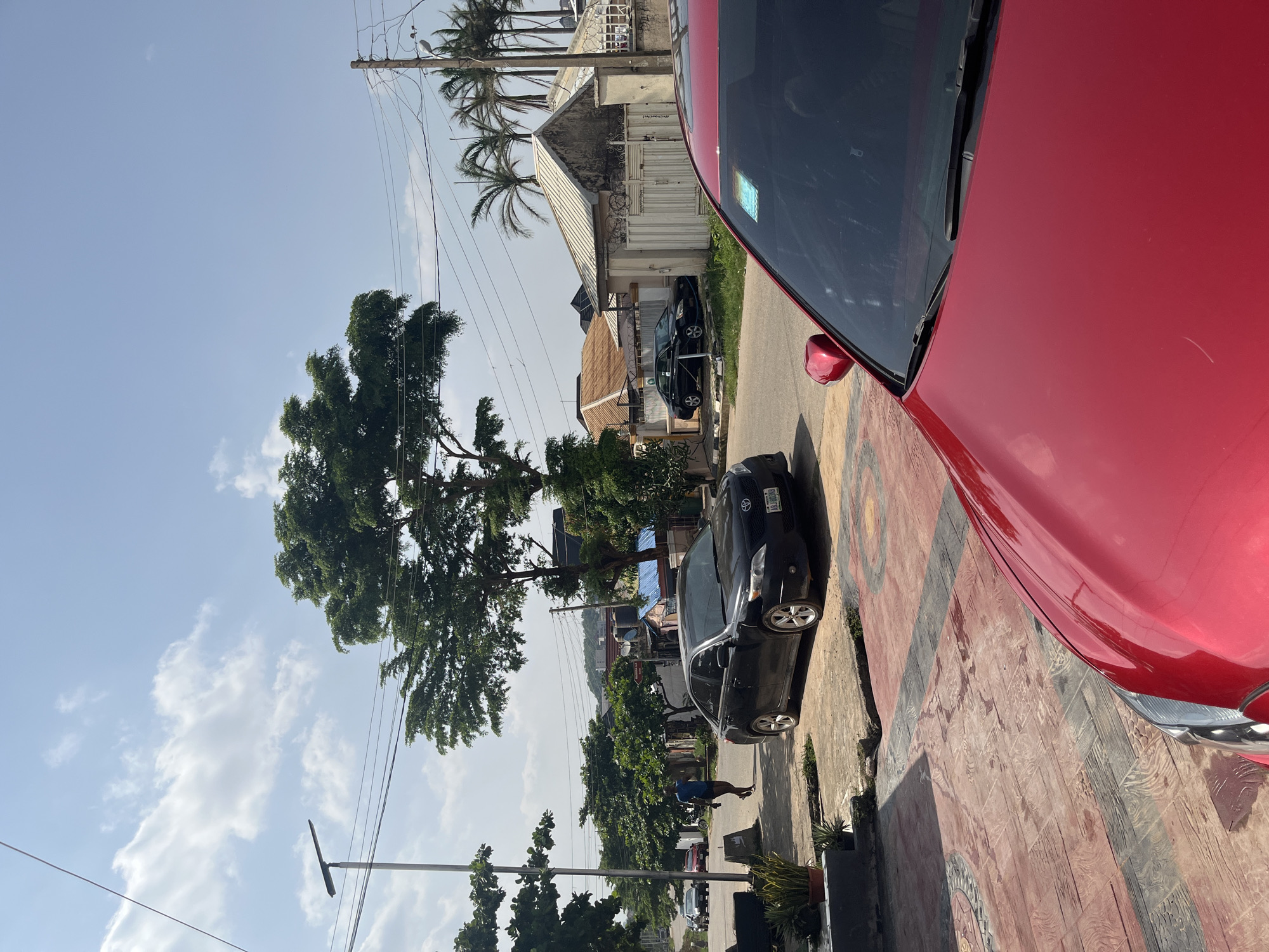 This image depicts a street scene in Abuja, Nigeria, under a bright, partly cloudy sky, suggesting it is daytime. The foreground is dominated by the vibrant red body of a car, its hood and windshield visible. To the left of the car, a black sedan is parked on the side of the road, and further down the street, another black car is visible. A person in a blue shirt is walking on the sidewalk.
The environment is characterized by lush green trees, including a large, mature tree with an expansive canopy. Palm trees are also present, adding to the tropical feel of the location. Buildings line the street, some appearing to be residential or commercial structures with corrugated metal roofs and decorative gates. Electrical wires are strung across utility poles, a common sight in many urban and suburban areas.
The sidewalk is paved with decorative tiles featuring geometric patterns in red, black, and grey. The overall impression is one of a well-established neighborhood or commercial area with a mix of residential and vehicular elements. The sunlight casts shadows, indicating a sunny day, and the presence of clouds suggests a dynamic weather pattern. The image captures a slice of daily life in Abuja, with cars, pedestrians, and the surrounding architecture contributing to the scene.