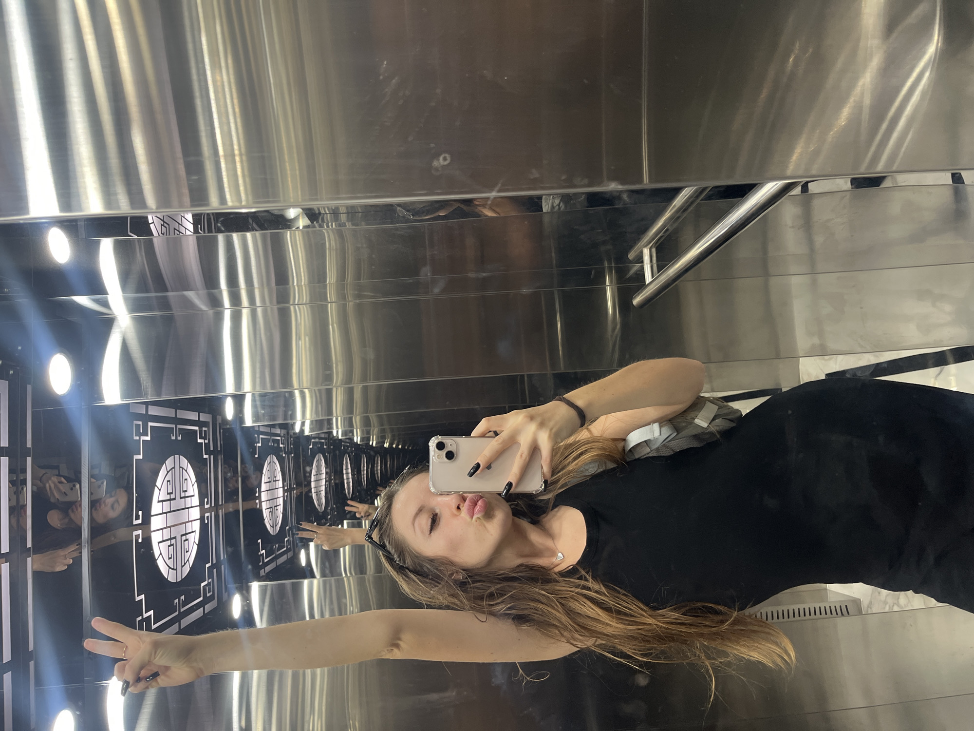 In an elevator, a young woman with long, light brown hair and dark nail polish poses for a selfie. She is wearing a black tank top and a silver necklace with a heart pendant. She holds a smartphone in her right hand and makes a kissing face while raising her left arm in a peace sign. The elevator walls are made of polished metal, reflecting the woman and the elevator's ceiling, which features a geometric design and lighting fixtures. The scene is set in an indoor environment, likely a building in Bac Nha Trang Ward, Vietnam, given the context of the provided information, although this specific detail is not visually evident in the image itself.