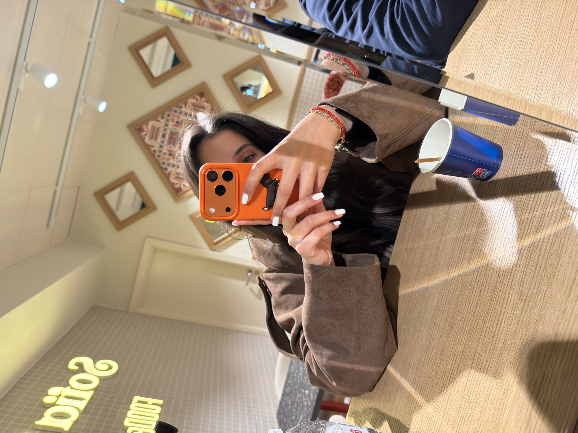 A person is captured in a mirror selfie, focusing on their reflection while holding an orange smartphone. The individual has long, dark hair, and her right eye is visible. She wears a brown jacket or coat and has white nail polish on her fingers. On her right wrist, she sports two bracelets, one red braided and one gold chain. The orange phone case features a black cat-like figure on the back.
The setting appears to be indoors, possibly a cafe or eatery. Reflected in the mirror is a light-colored wall adorned with several small, square frames, some containing mirrors and one displaying a patterned textile or artwork. Overhead, track lighting with white spotlights illuminates the space. A wooden table or counter surface is prominent in the foreground and to the right, where an empty blue paper cup with a wooden stir stick rests. Part of another person, wearing dark blue pants, is reflected above the main subject. In the lower left, a glowing yellow neon sign is reflected and reversed, appearing to spell "SOOOS" and "SPOOF," likely indicating "SOFIA FOODS" or a similar establishment name. A partially visible water bottle is also near the bottom edge.