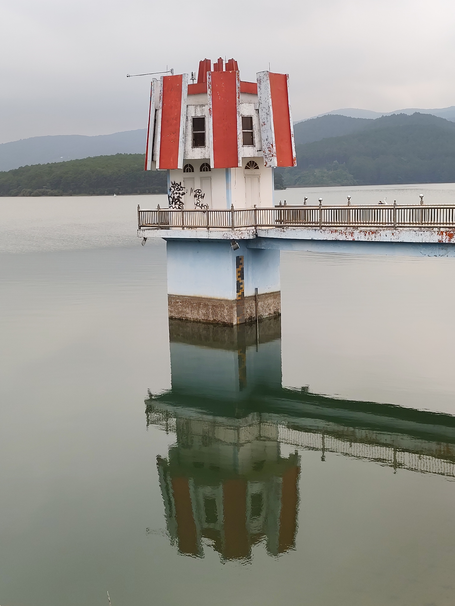 The image depicts a structure resembling a lighthouse or water tower located in Tuyen Lam Lake, Da Lat, Vietnam. The tower has a white base and a red and white upper section with windows. Graffiti is visible on the white section beneath the upper structure. A bridge extends from the tower, featuring a railing on both sides. The structure is reflected in the calm water of the lake. In the background, tree-covered hills fade into a hazy sky. A water level gauge is attached to the side of the tower support.