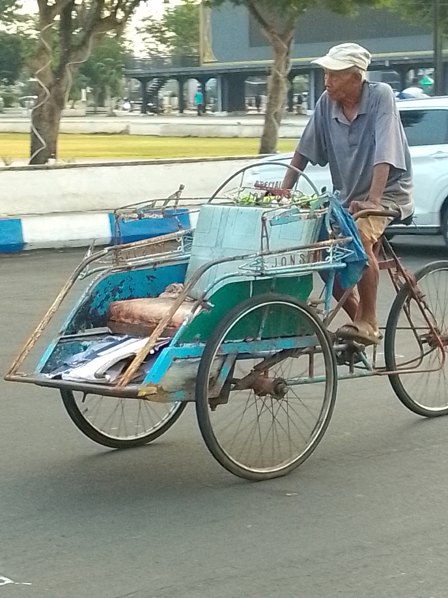 An elderly Indonesian man wearing a light-colored cap and a grey polo shirt pedals a three-wheeled cycle rickshaw. The rickshaw has a passenger compartment with a blue and green exterior and appears to be loaded with various items, including what looks like a brown cushion and some folded materials. The man is seated on the rickshaw, with his legs positioned to pedal. The vehicle is on an asphalt road, with a white and blue curb to the left of the image. In the background, trees and a building with an arched structure are visible, suggesting an outdoor, urban setting. The scene appears to be in the city of Jember, Indonesia, based on the provided context.
