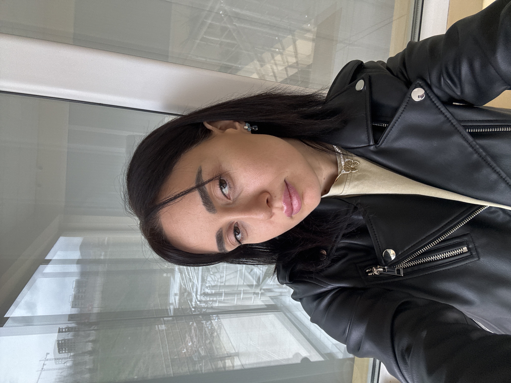 A female individual is captured in a close-up, chest-up selfie. She has dark, shoulder-length hair and is looking directly at the camera with a neutral expression. Her lips are adorned with pink lipstick. She wears a black leather-style moto jacket featuring an asymmetrical zipper, snap-button lapels, and zippered pockets. Beneath the jacket, a light beige/cream-colored top is visible. A small earring with blue and white stones is worn on her right ear, and a silver-colored necklace with a butterfly pendant is visible around her neck.
The subject is indoors, positioned in front of a large, multi-pane window with white frames. Through the window, an overcast sky is visible over a distant landscape dominated by gently sloping, partially vegetated hills. The background reveals active construction, including multiple unfinished building frameworks and at least one tall tower crane. Several completed multi-story residential or commercial buildings are also present further in the distance. A small, red octagonal sign, possibly a stop sign, is faintly visible on a distant road.
This scene is located in Tbilisi, Georgia, specifically in a developing area characterized by modern residential construction on hilly terrain, such as the vicinity of Lisi Lake.