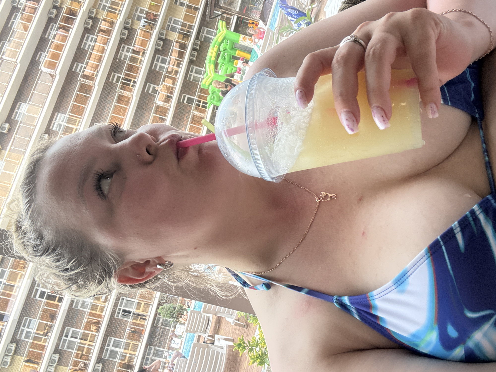 A young woman with light skin and blonde hair is depicted drinking from a clear plastic cup filled with a yellow, slushy-like beverage through a pink straw. She is wearing a blue and white patterned bikini top and a gold necklace with a small animal-shaped pendant. A silver ring adorns a finger on her right hand, which is holding the cup, and her nails are painted light with tiny pink specks. She also has a small nose piercing and an earring in her right ear.
The image is captured outdoors during daytime, under bright sunlight, suggesting warm weather. In the background, a large, multi-story building with numerous windows, balconies, and air conditioning units is visible, likely a hotel or apartment complex. Below the building, parts of a resort or pool area can be seen, including white lounge chairs, a swimming pool, and a green inflatable structure, possibly a bouncy castle or slide. The overall scene conveys a relaxed, leisure setting, perhaps at a vacation destination. No discernible text is visible in the image.