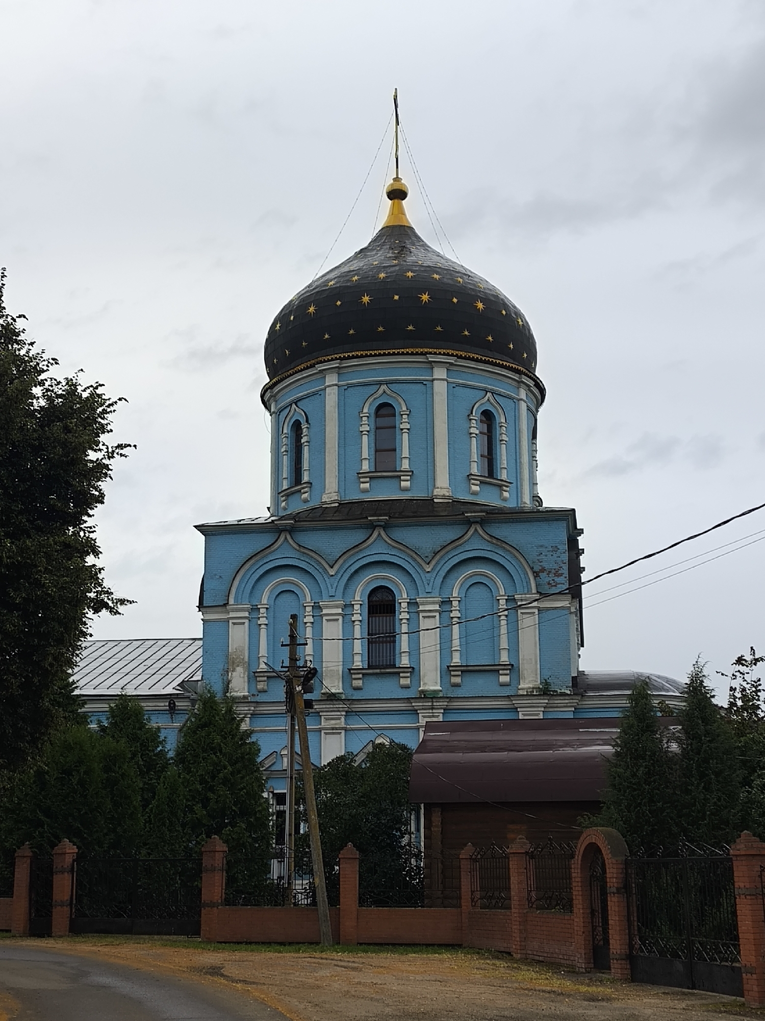This is a full-frame, eye-level, outdoor, medium shot photograph of a blue church with a dark dome in the city of Pokrov, Russia. The dome is decorated with gold stars and topped with a golden cross. The church has white decorative trim around its windows and architectural features. In the foreground, a dark asphalt road curves towards the left side of the frame. A low, red brick fence with black metal gates separates the road from the church grounds. Evergreen trees and a dark-roofed building are visible behind the fence and to the right of the church. The sky is overcast and gray.