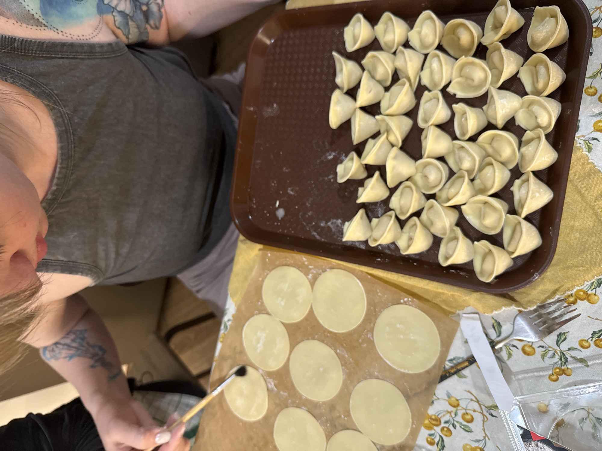 A person is making tortellini. The individual is pictured from above, wearing a gray tank top and shorts, with tattoos visible on their arm and shoulder. A brown tray filled with completed tortellini sits on a yellow cloth. A sheet of parchment paper with several round dough pieces is also visible. The person is using a brush to apply something to the dough. A fork and packaging material are on a floral patterned tablecloth. The location is unknown and there is no evidence in this image to suggest the city of Krasnoyarsk, Russia.